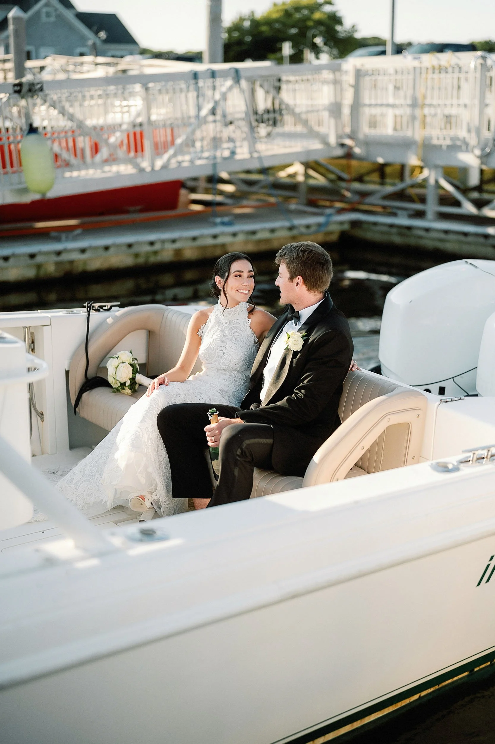 Bride and groom sitting together on a boat, smiling at each other, near a dock with boats and water in the background.