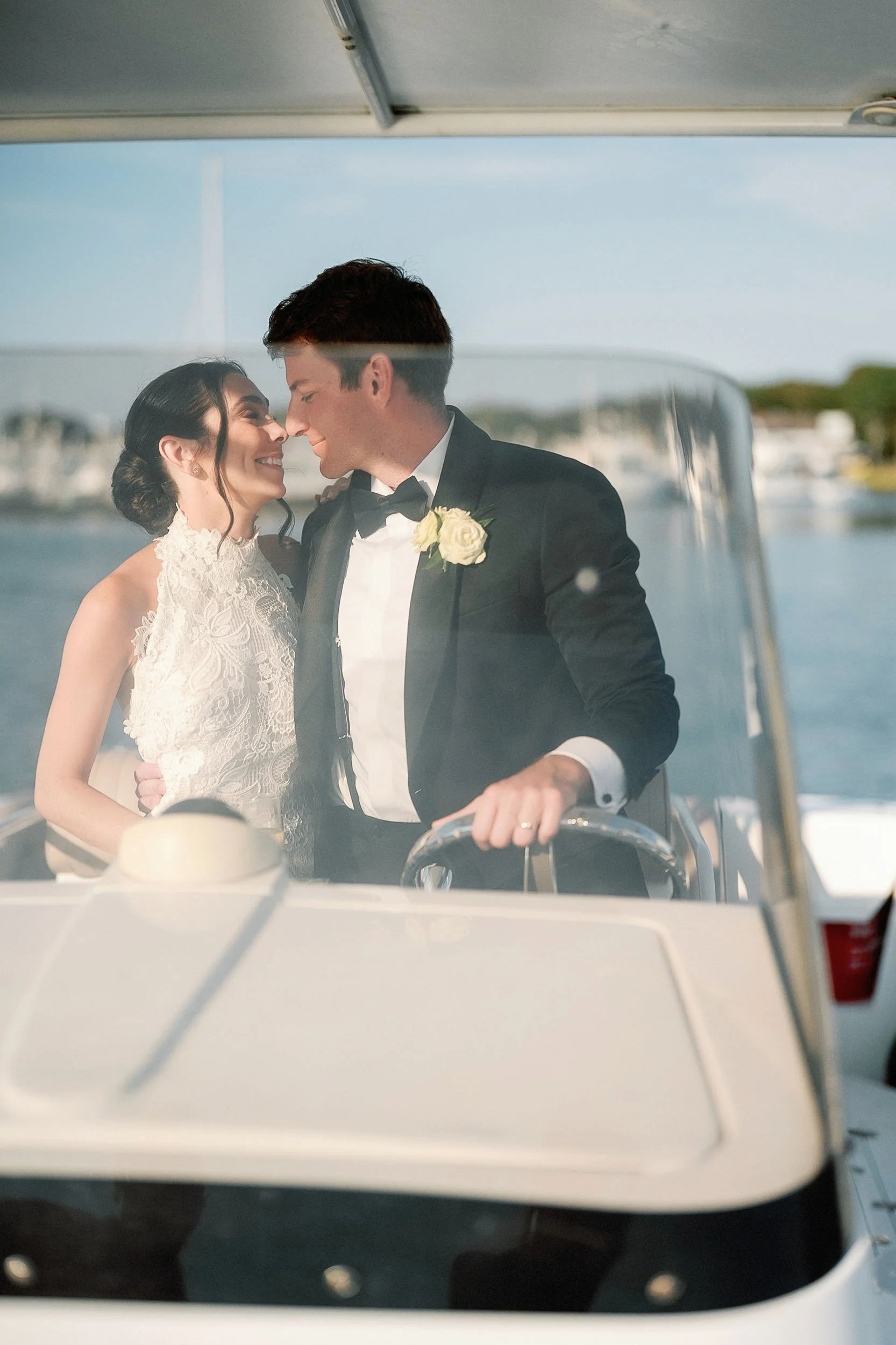 A bride and groom in wedding attire on a boat, smiling and about to kiss, with water and buildings in the background.