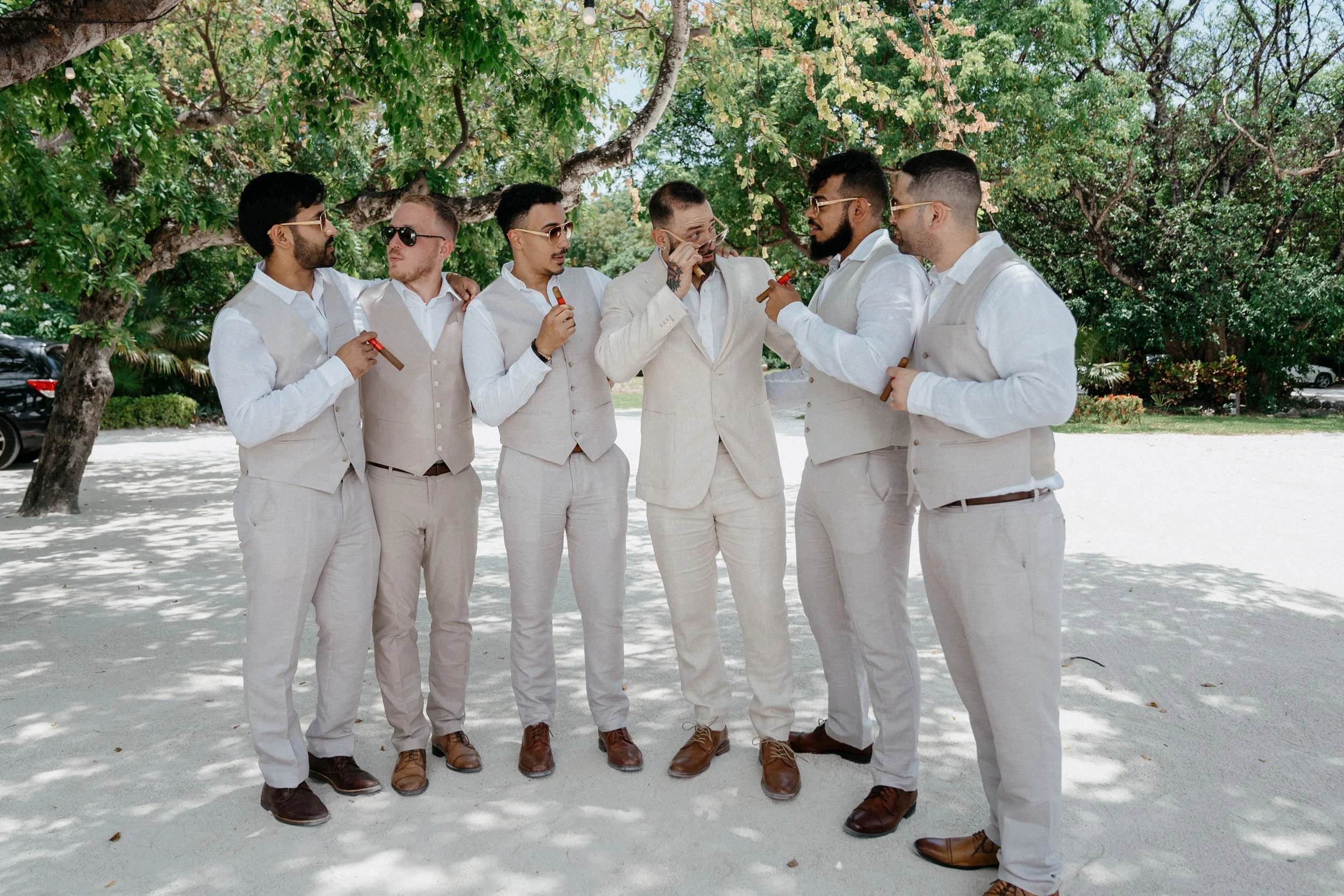 A group of seven men dressed in light-colored suits and sunglasses standing outdoors under a tree, holding cigars, engaged in conversation.