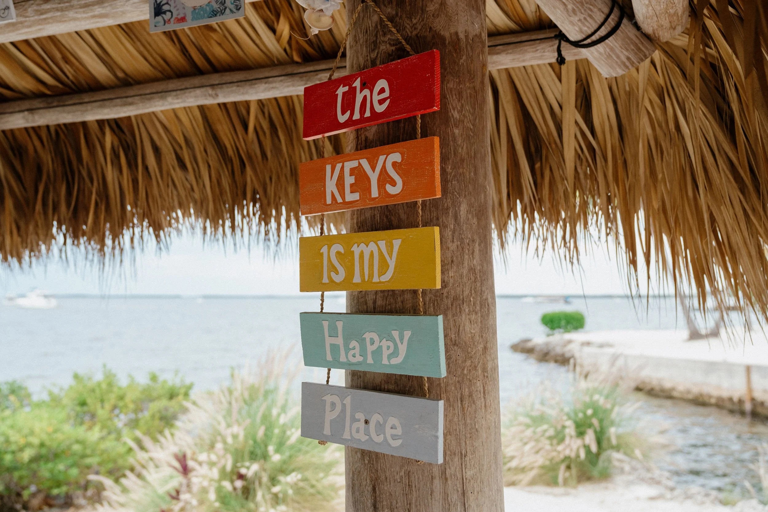Colorful wooden sign hanging from a thatched roof pole reading "the KEYS is my HAPPY Place" in a beach setting with water and boats in the background.