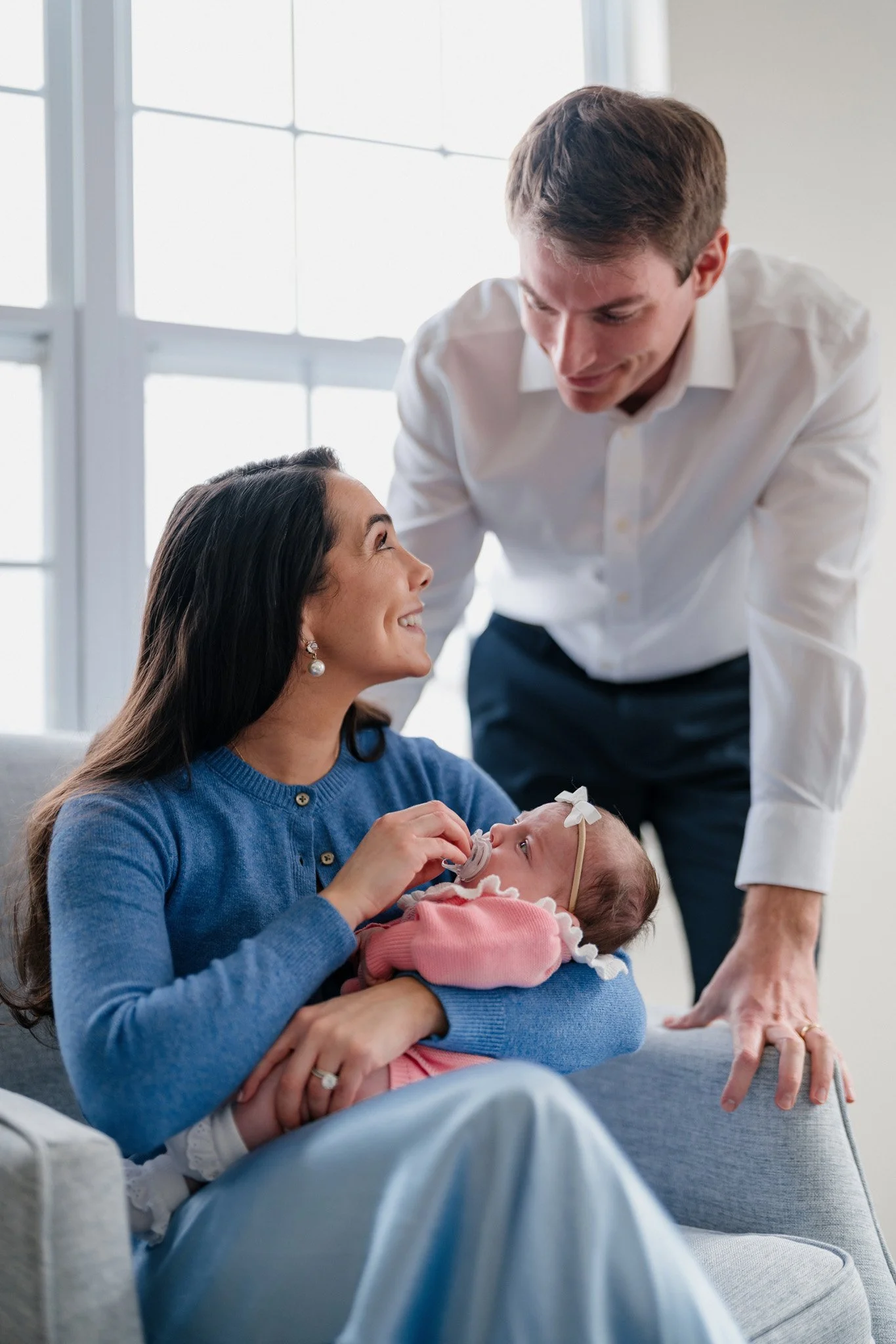 A woman holding a baby with a pacifier, smiling at a man leaning over her, in a bright room with large windows.