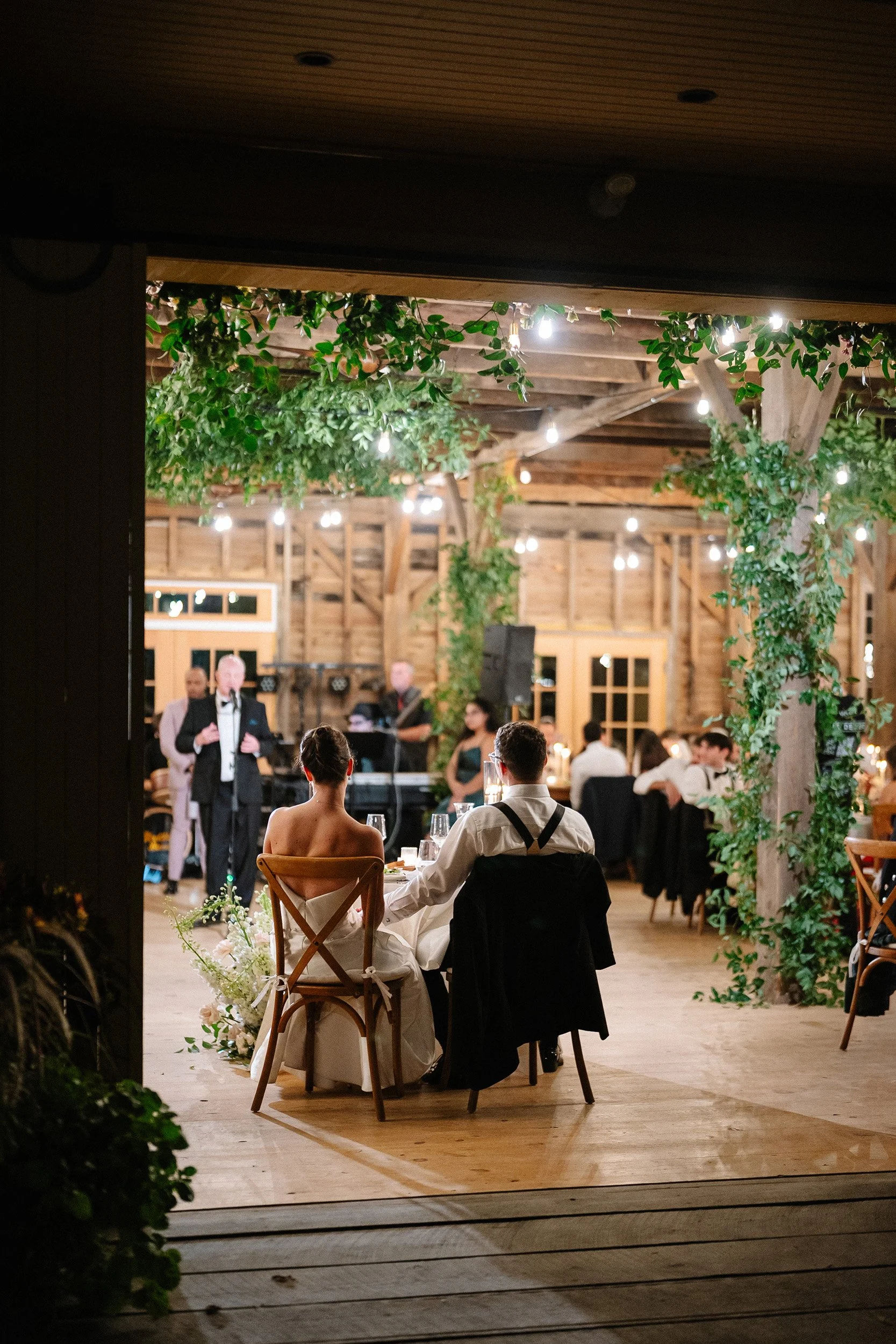 A newlywed couple sitting at a table in a rustic barn decorated with greenery and string lights, during their wedding reception.