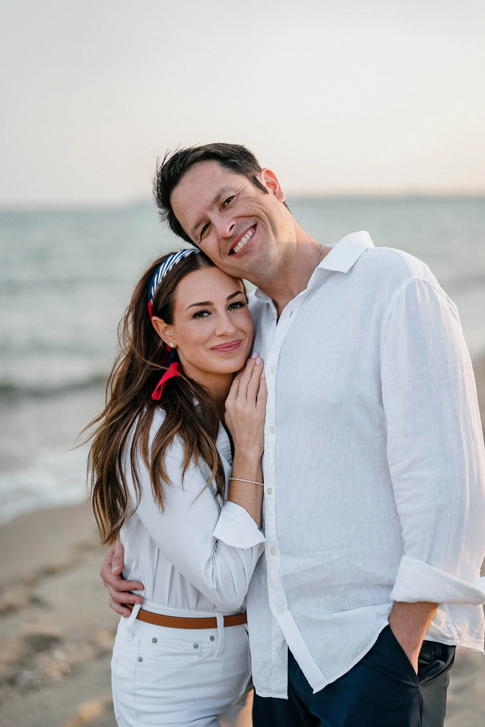 A smiling man and woman hugging at the beach during sunset, dressed in white and casual clothing.