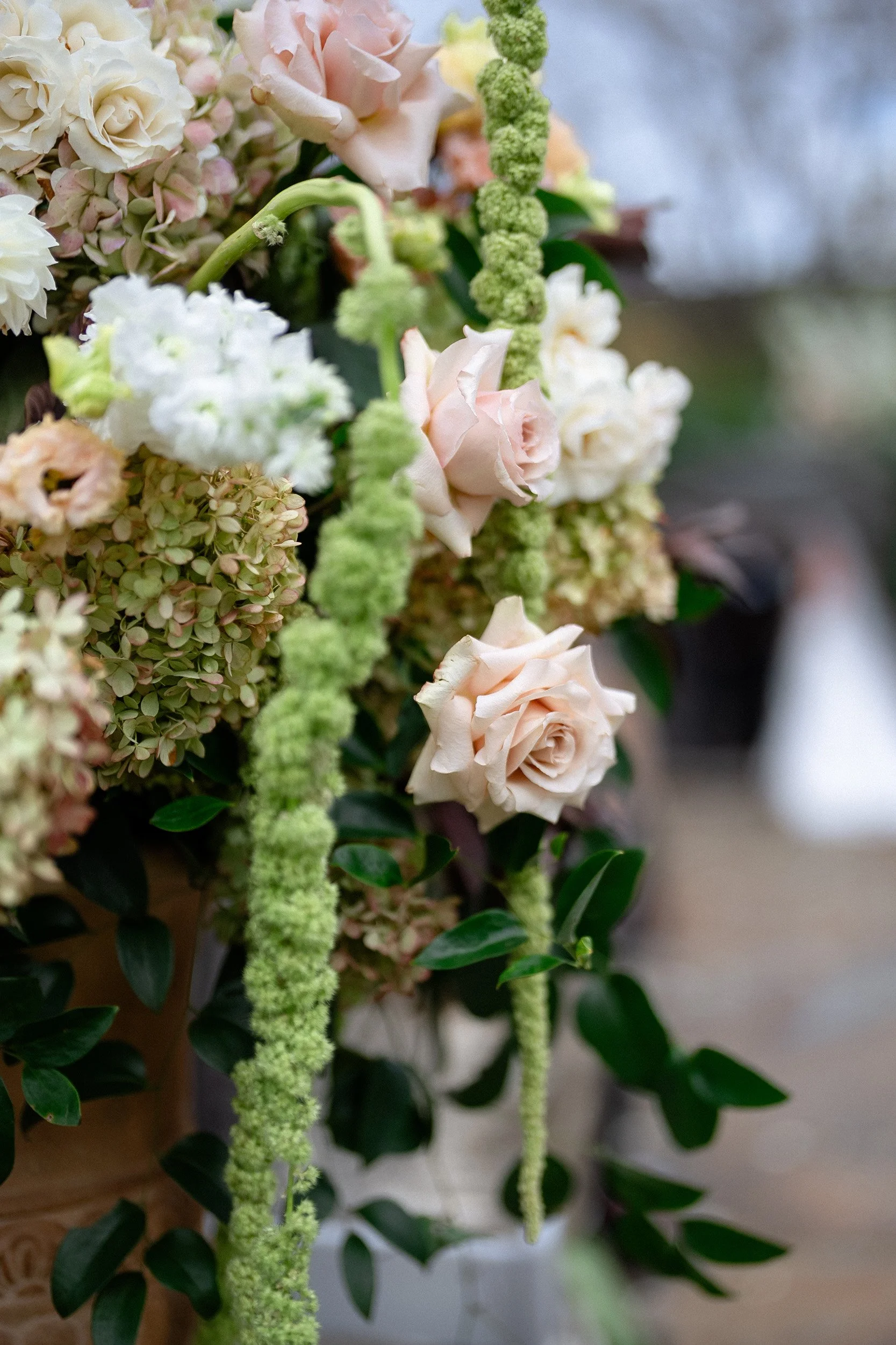 A floral arrangement with pale pink roses, green hanging amaranthus, white hydrangeas, and greenery, set against a blurred outdoor background.