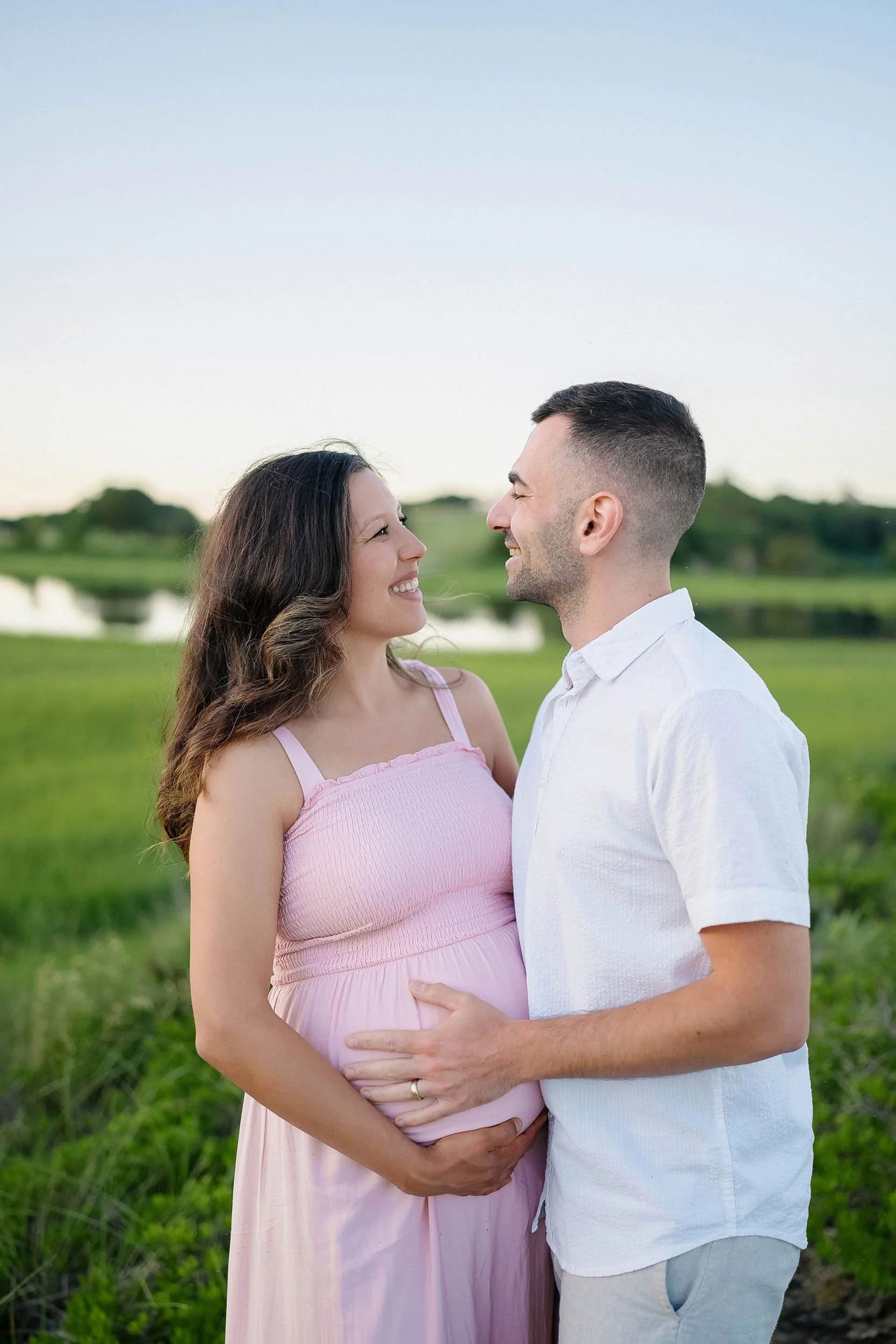 A couple standing outdoors, smiling at each other, with the woman pregnant, holding her belly, in a green field near a body of water during sunset or early evening.