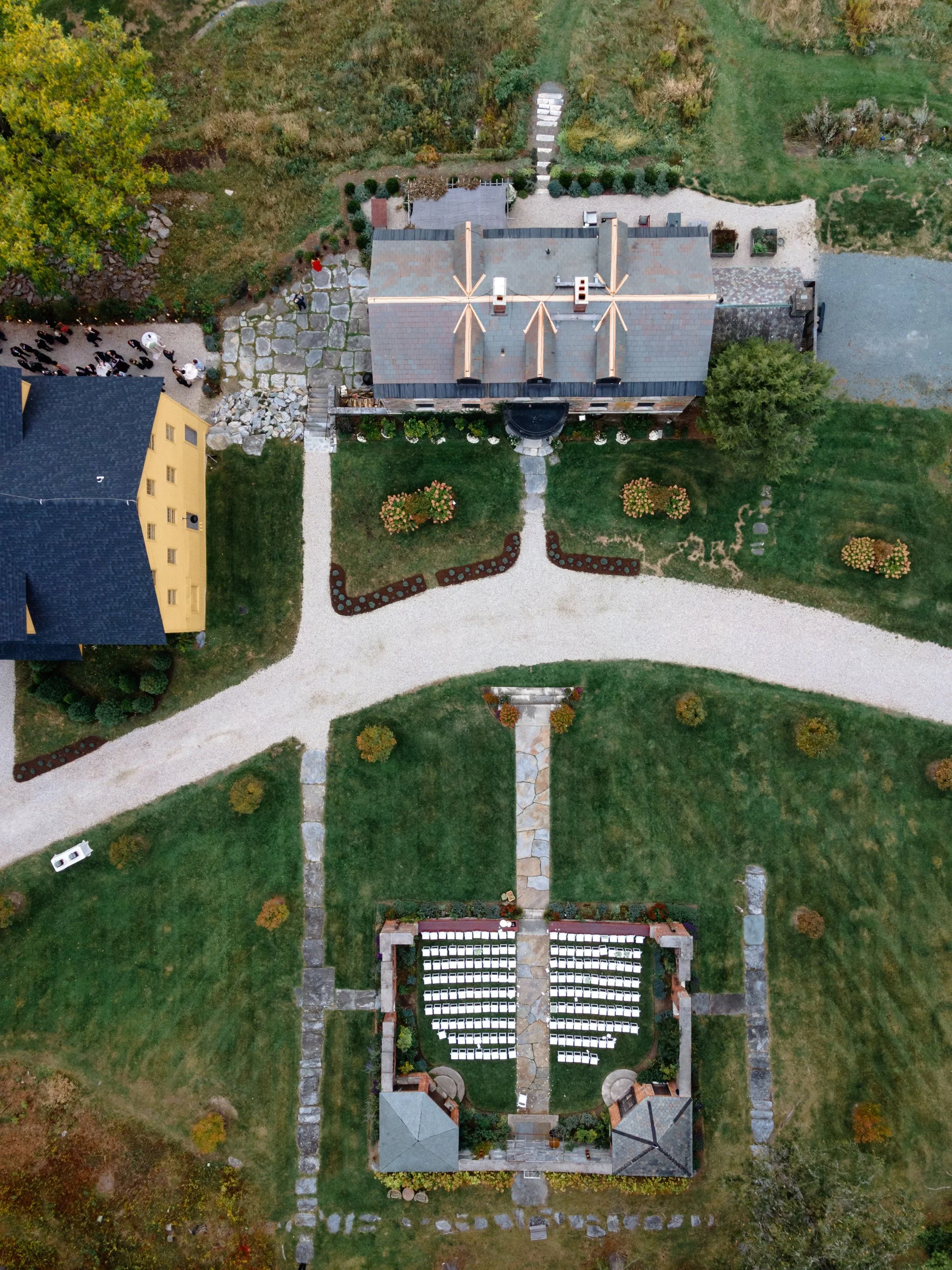 Aerial view of a wedding ceremony setup with rows of chairs on a green lawn, a pathway leading to a small stage, and a historic building in the background with a gravel driveway and surrounding gardens.