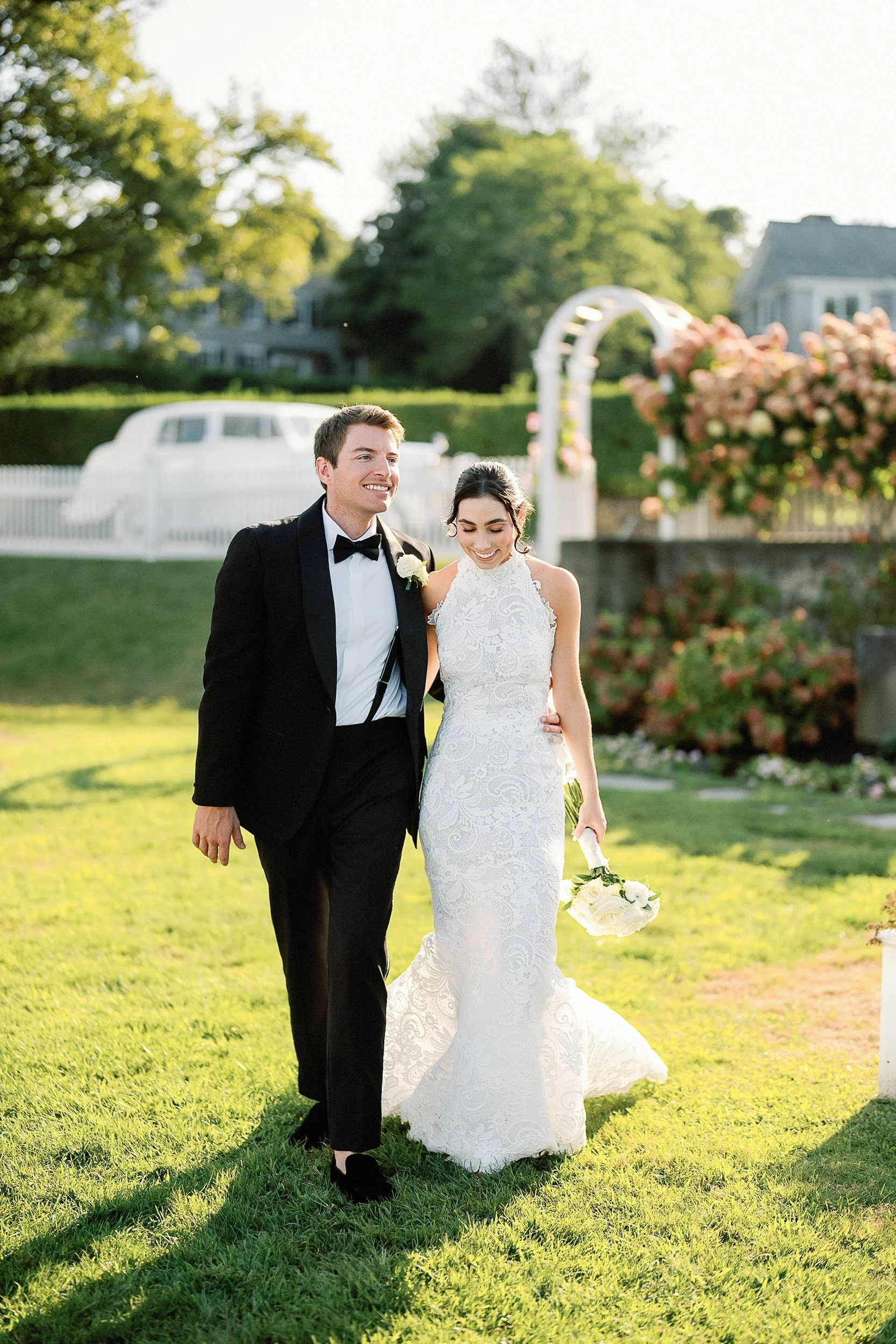 Bride and groom walking outdoors during wedding ceremony, smiling and happy, with trees and flowers in the background.