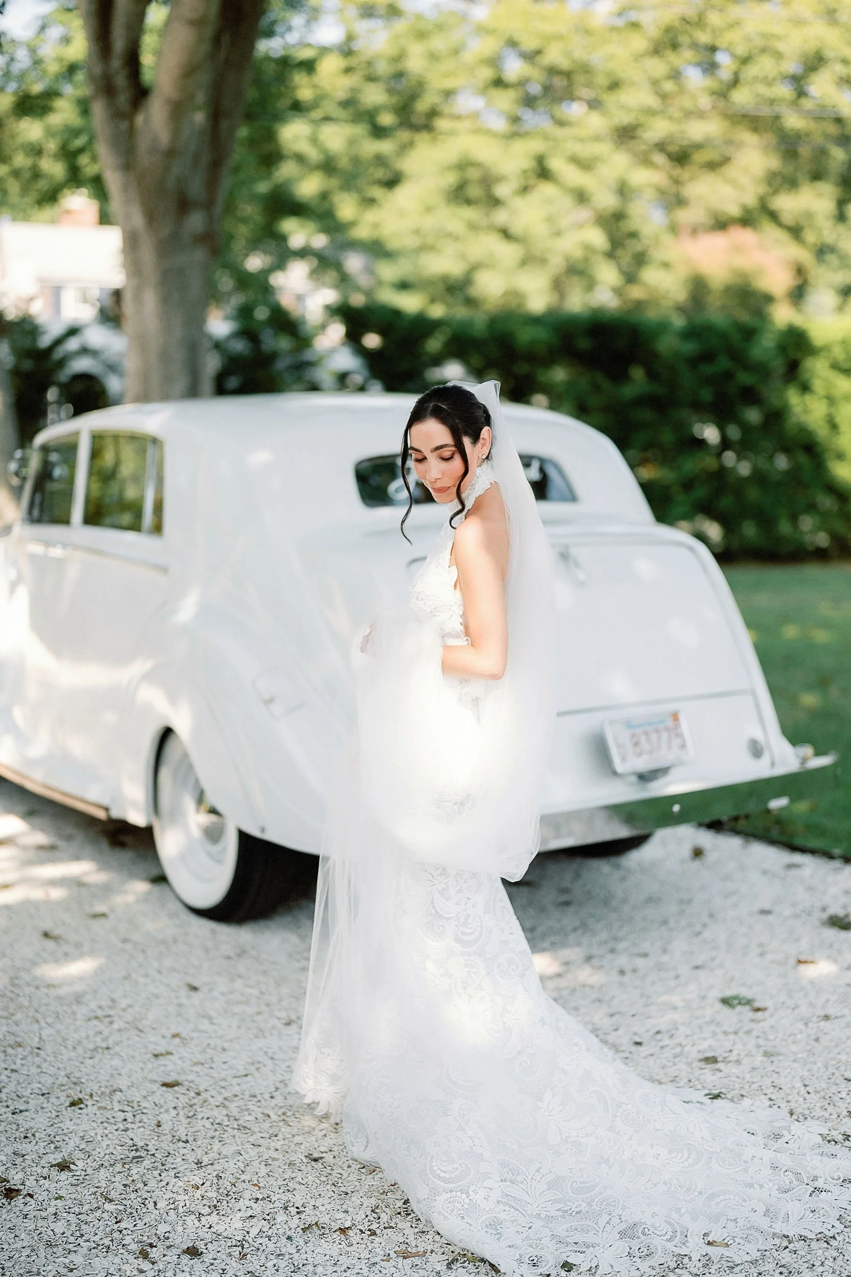 A bride in a white lace wedding gown and veil stands outdoors near a vintage white car, with green trees and a sunny sky in the background.