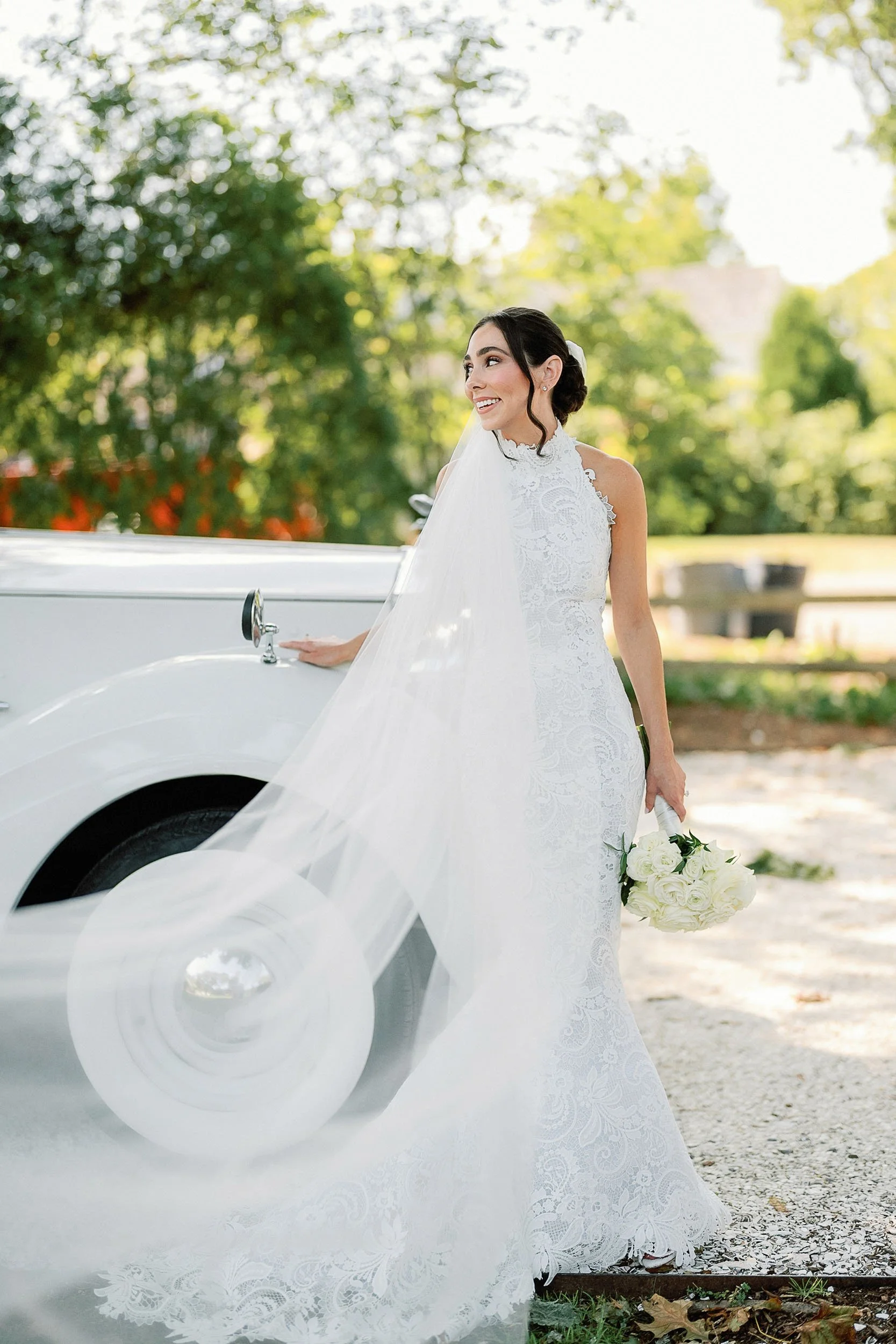 Bride in a white lace wedding dress holding a bouquet of white roses stands outdoors next to a vintage white car, with green trees and a sunny sky in the background.