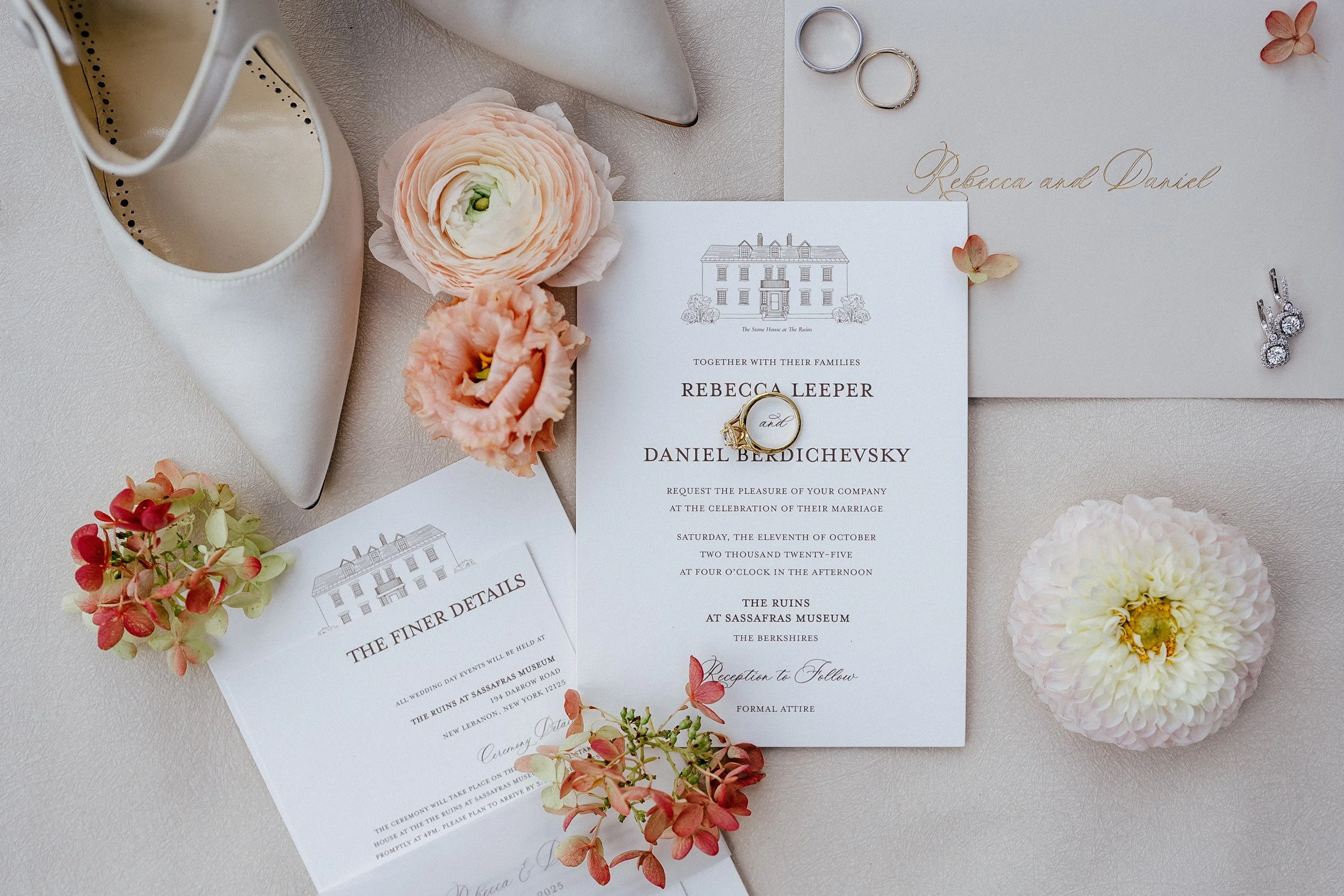 Wedding invitation with rings, flowers, and an ivory shoe, decorated with pink and white blossoms on a white surface.