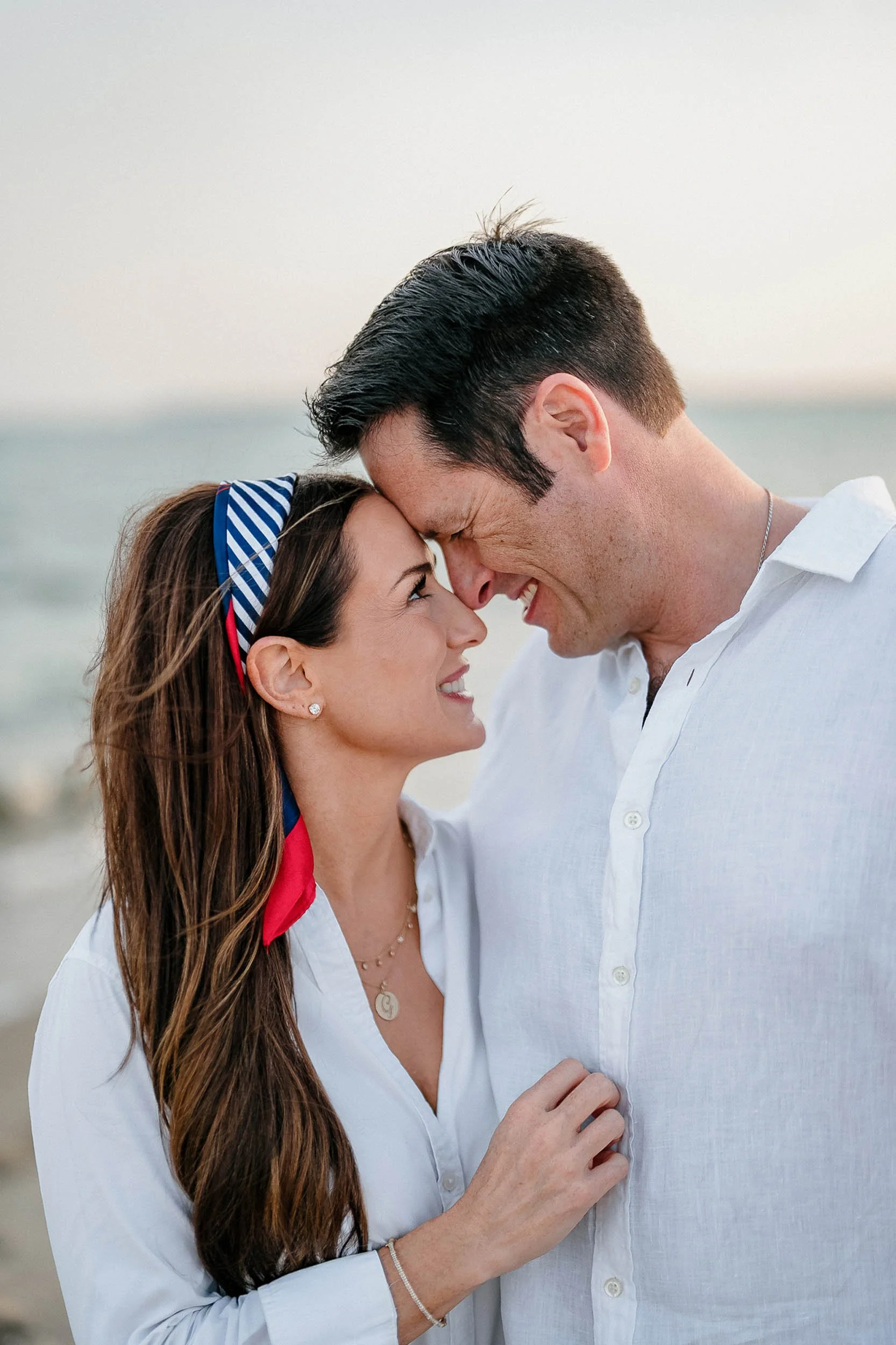 A couple at the beach, touching foreheads and smiling, with ocean in the background.