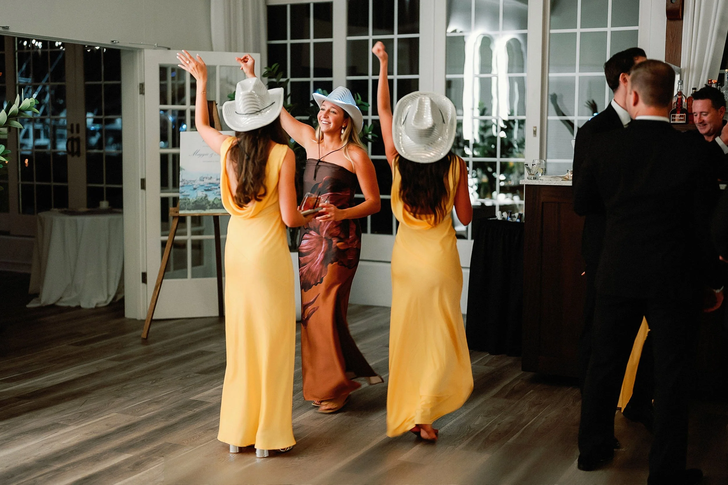 People dancing and socializing at a party or celebration in an indoor venue with large windows and wooden flooring.