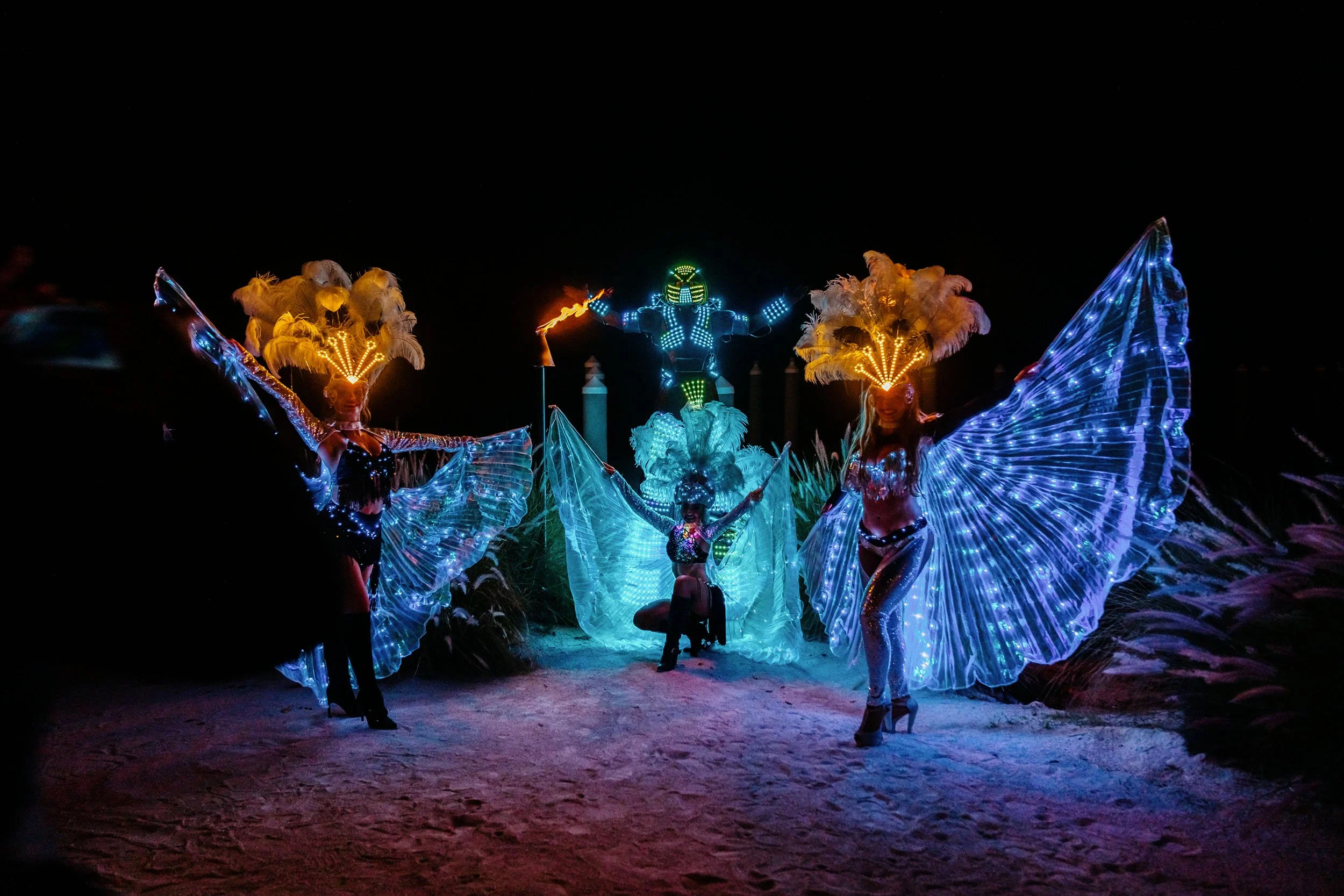 Night scene of performers in illuminated, feathered costumes with large wings, posed in front of a figure resembling a robot with LED lights and a flame, against a dark background.