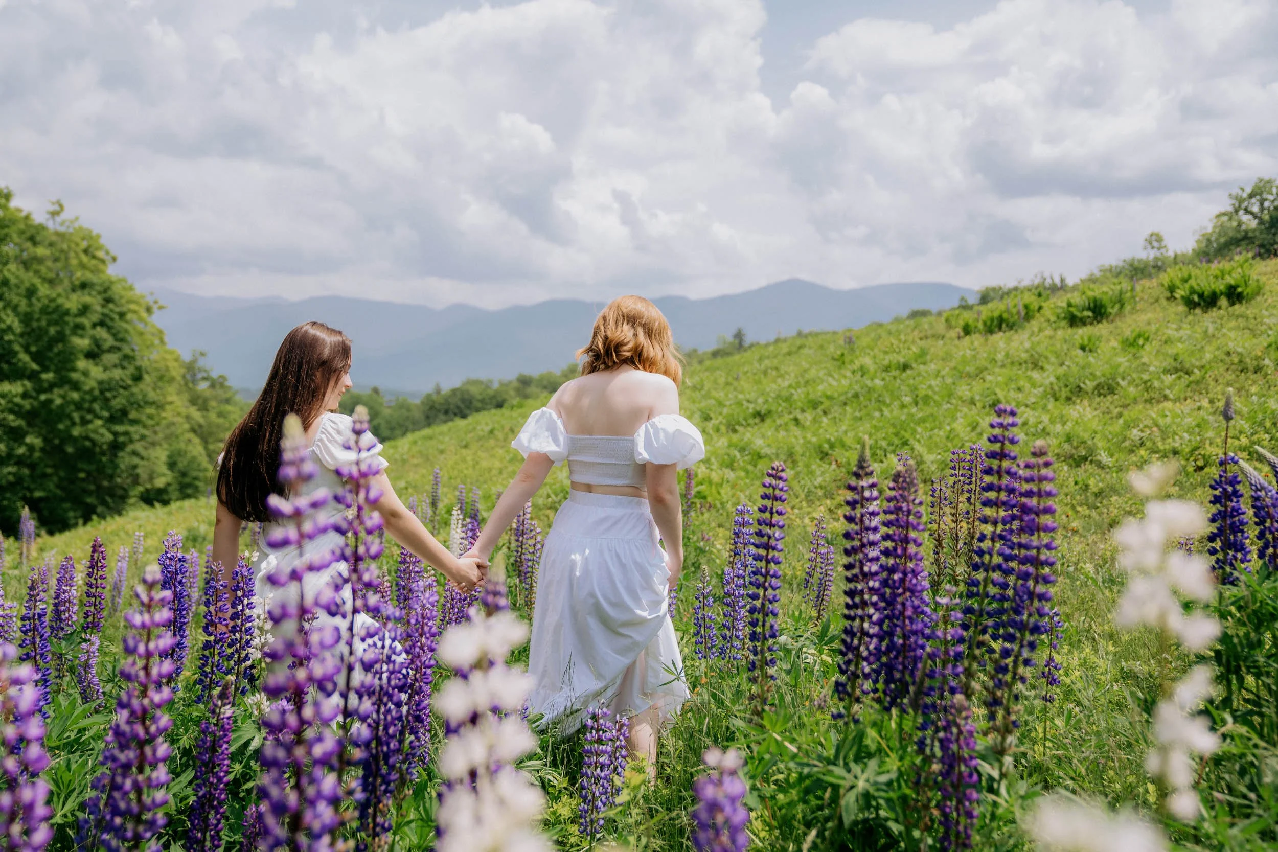 Two women in white dresses holding hands and walking through a field of purple and white flowers on a cloudy day with mountains in the distance.