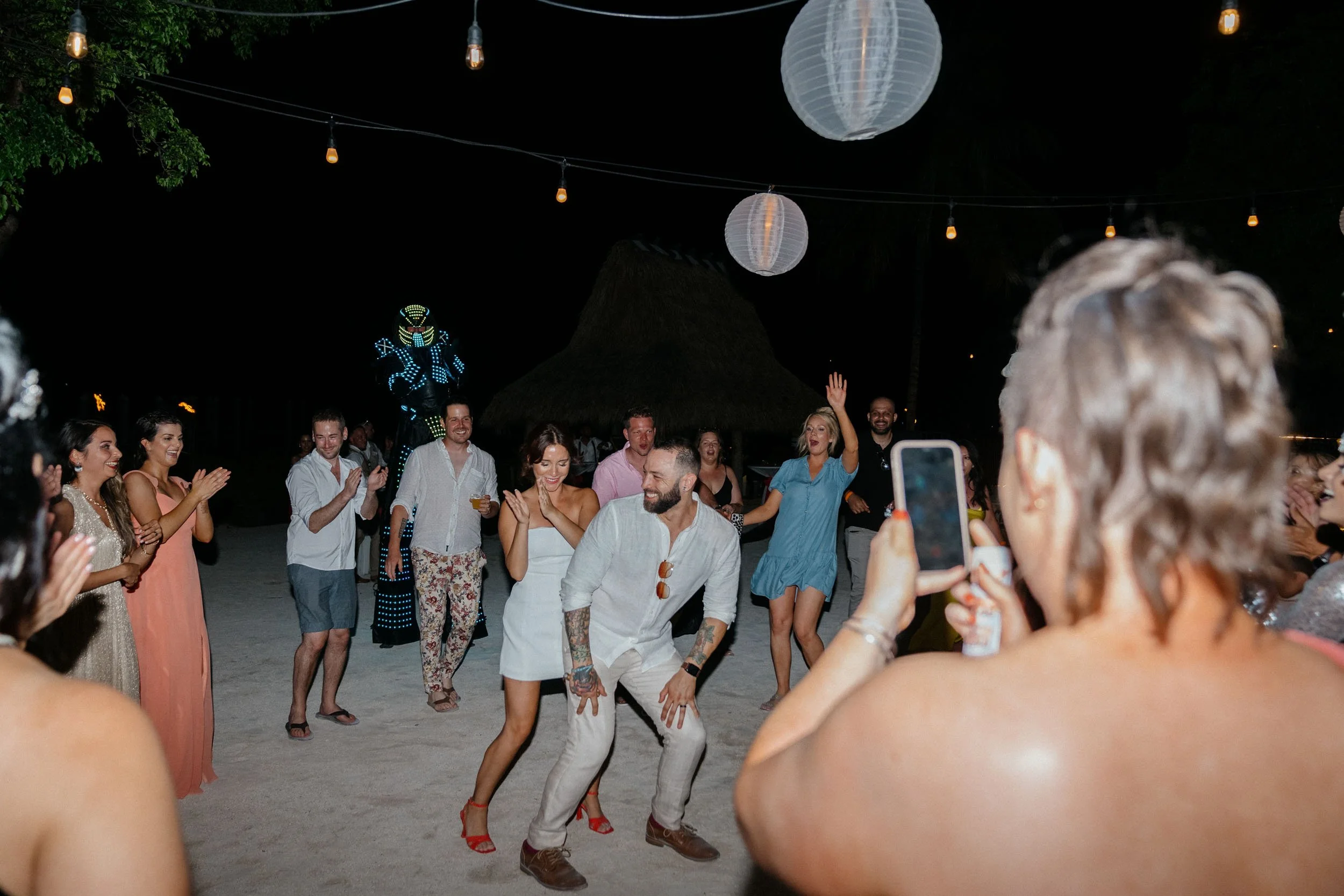 People dancing and having fun at an outdoor nighttime party, some holding drinks, with string lights and paper lanterns overhead, and a decorative lit figure in the background.