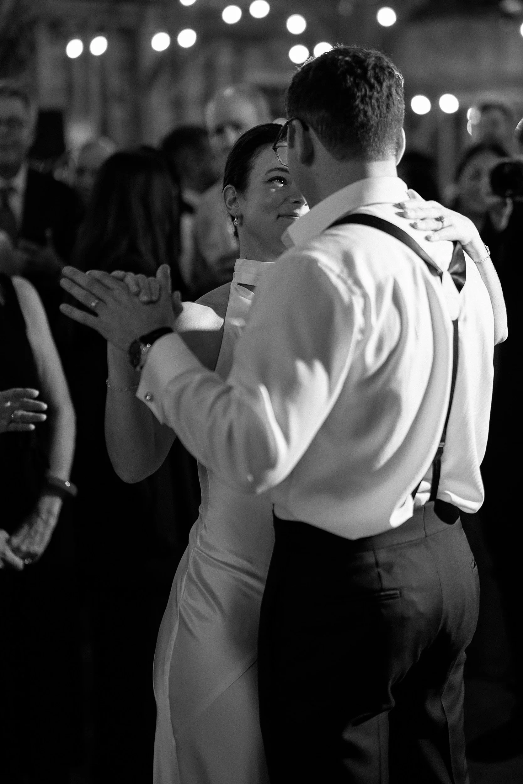 A black-and-white photo of a couple dancing closely at a wedding reception, surrounded by guests, with string lights hanging overhead.