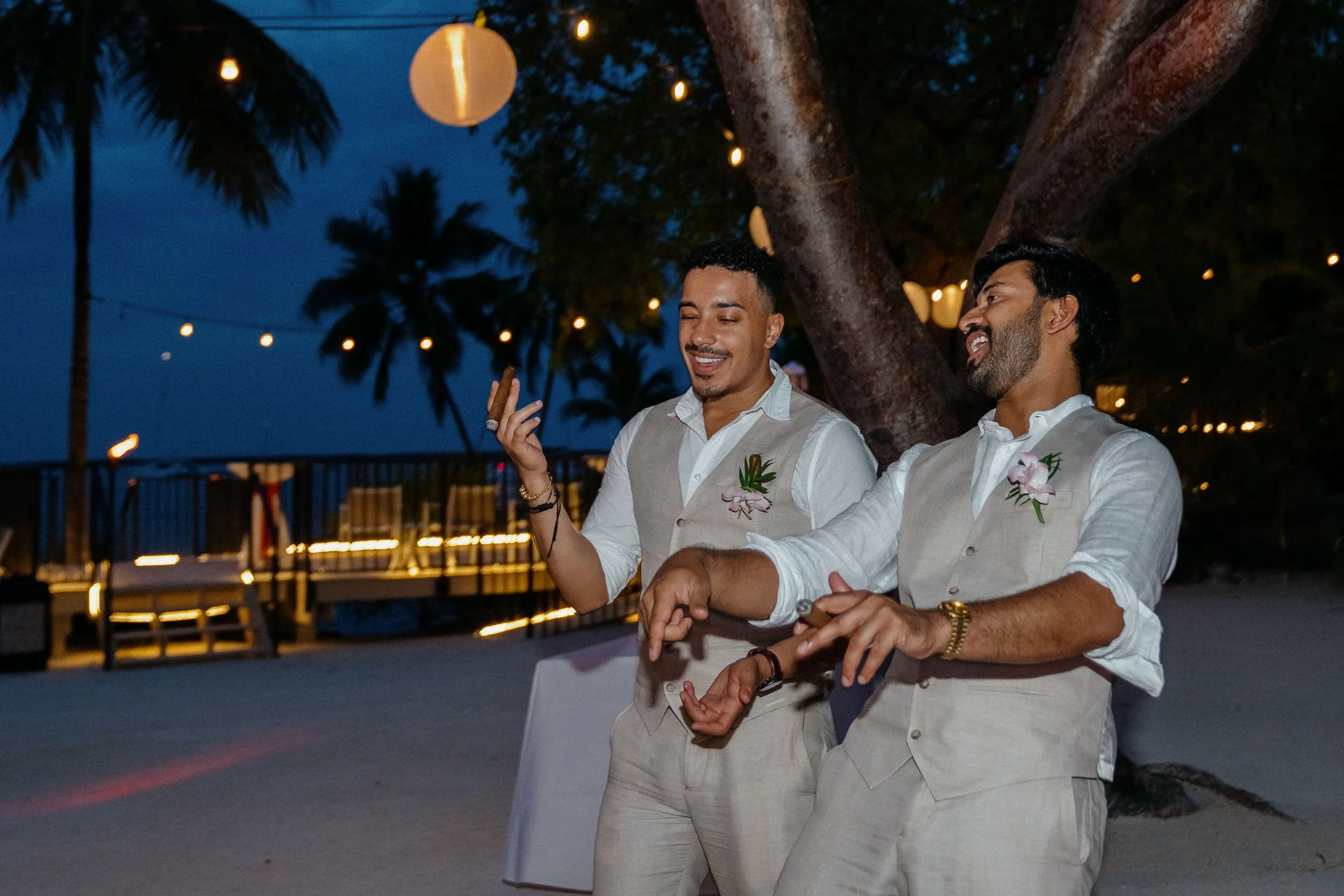 Two men dressed in light-colored vests and shirts, smiling and dancing outdoors at night during a beach wedding, with string lights and lanterns hanging from trees, palm trees in the background.