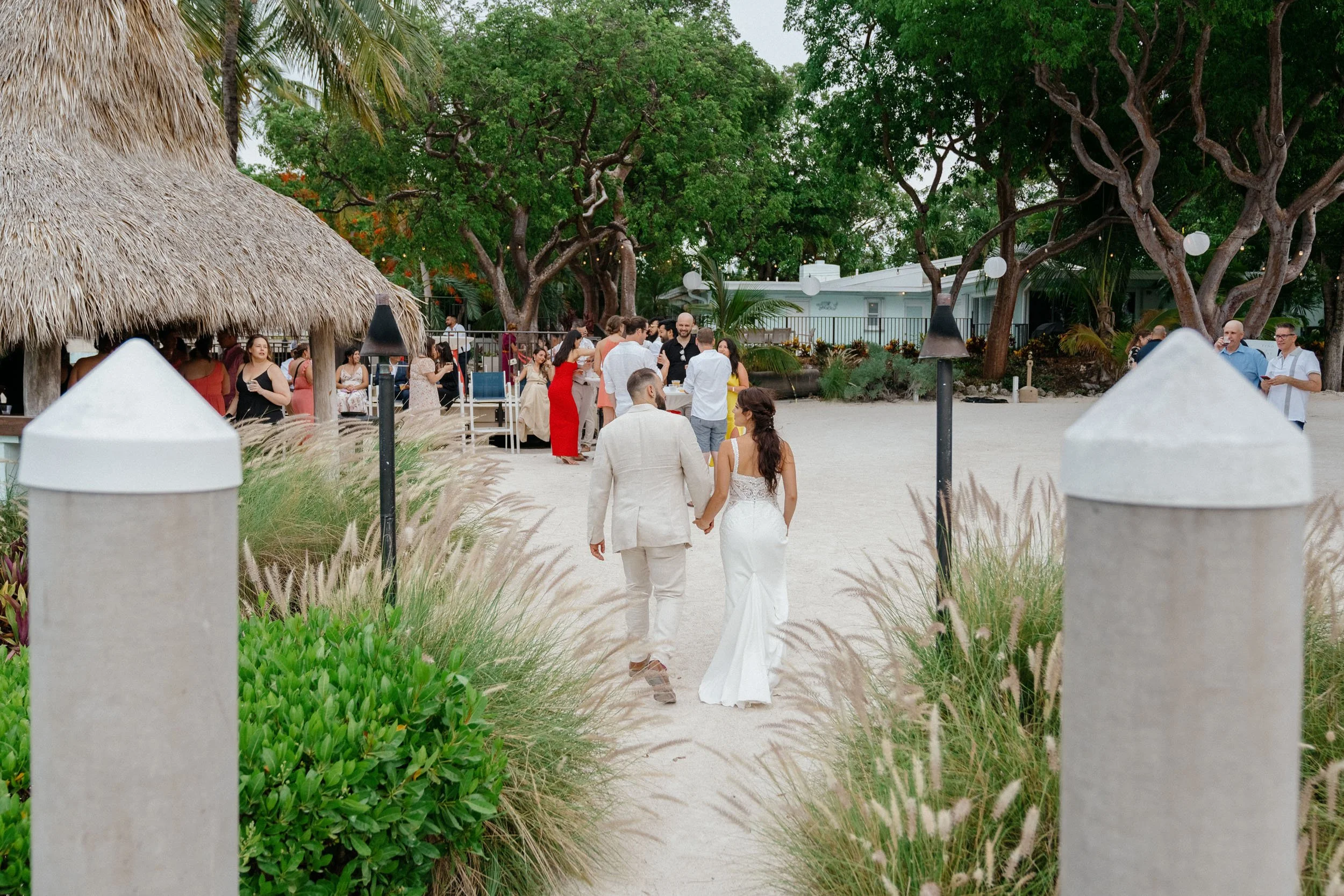 A newlywed couple holding hands walking towards a beachside outdoor reception with guests, trees, and a thatched roof hut in the background.