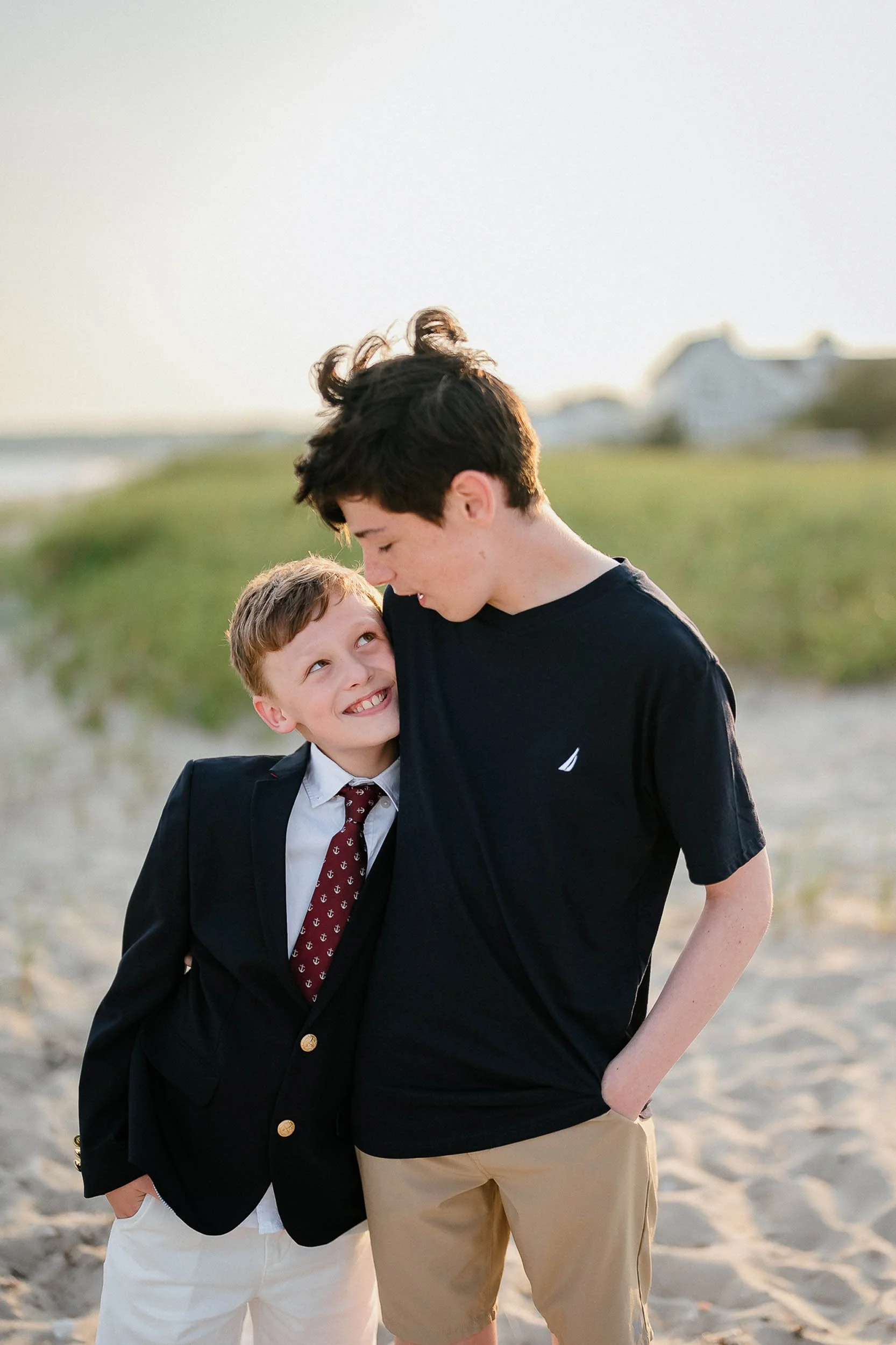 Two boys on the beach, one in a suit and the other in a t-shirt, smiling and looking at each other.