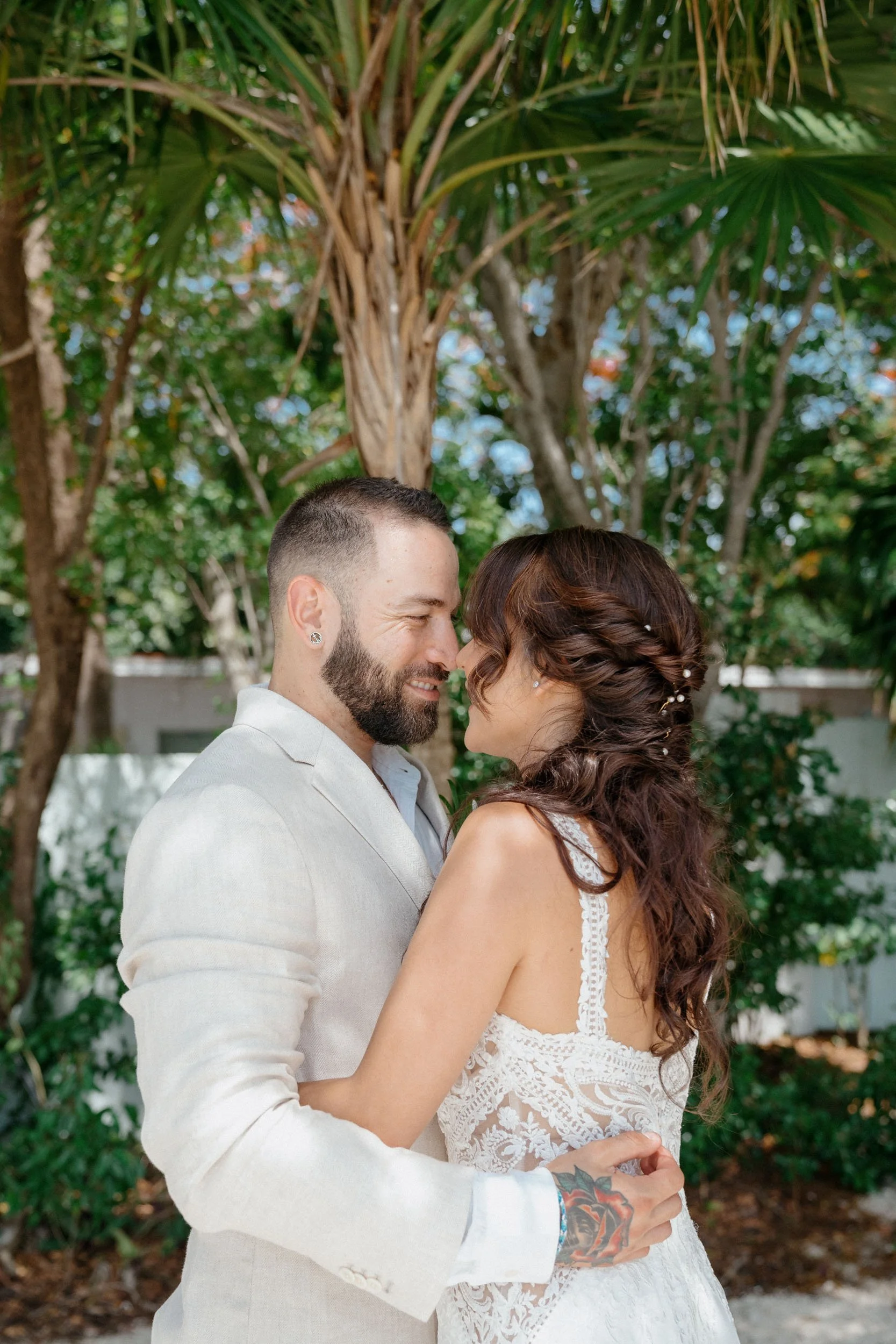 A couple in wedding attire embracing outdoors beneath trees, facing each other with foreheads touching.