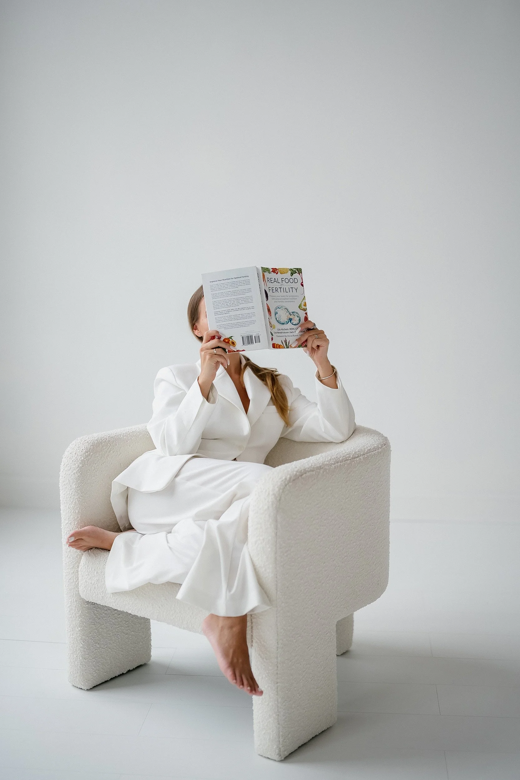 A woman in a white suit is sitting on a textured white armchair, with one leg crossed over the other, reading a book titled 'Real Food Fertility' in a minimalistic, white-room setting.
