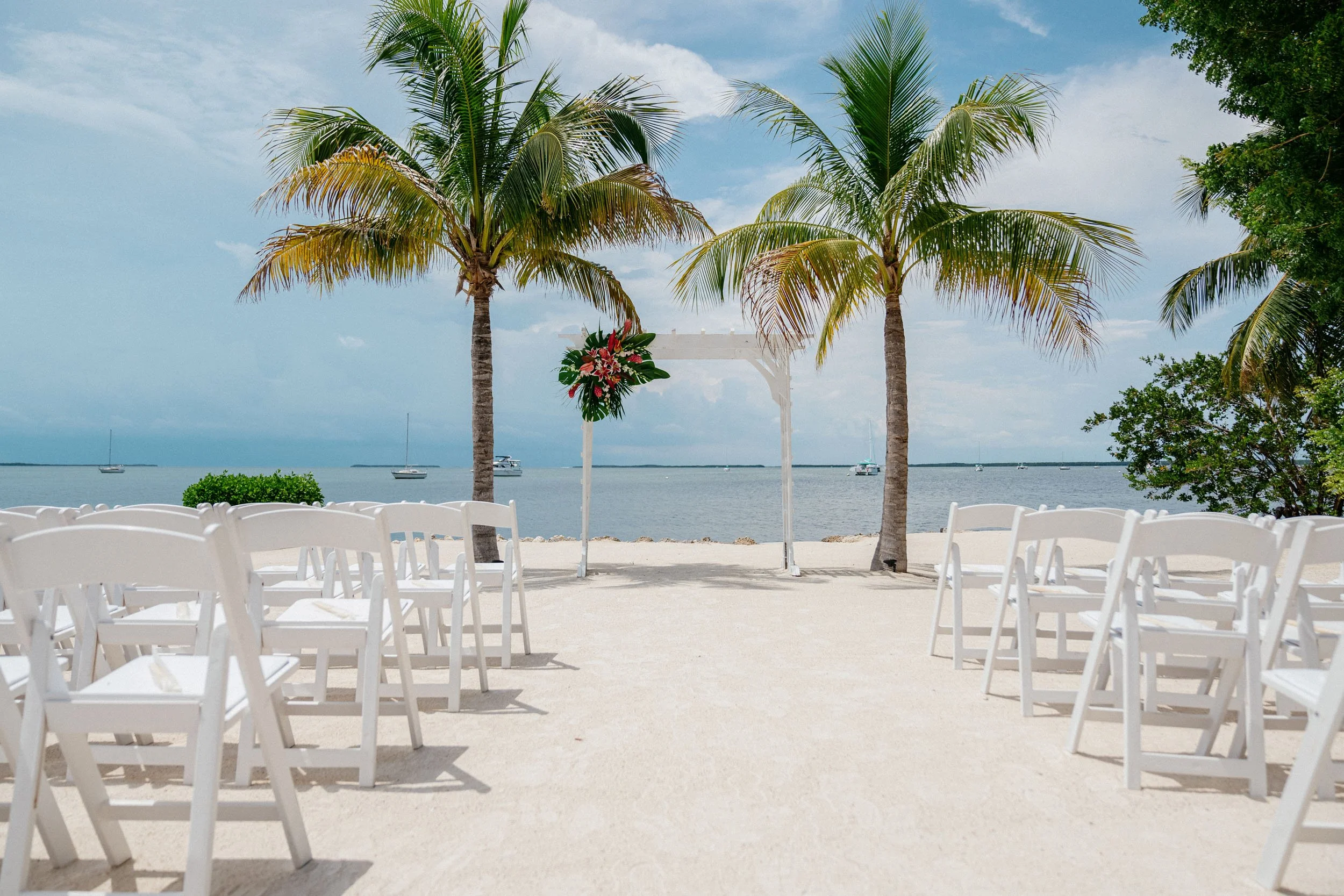 Beach wedding setup with white chairs, a decorated arch, palm trees, and a view of the ocean with boats in the distance.