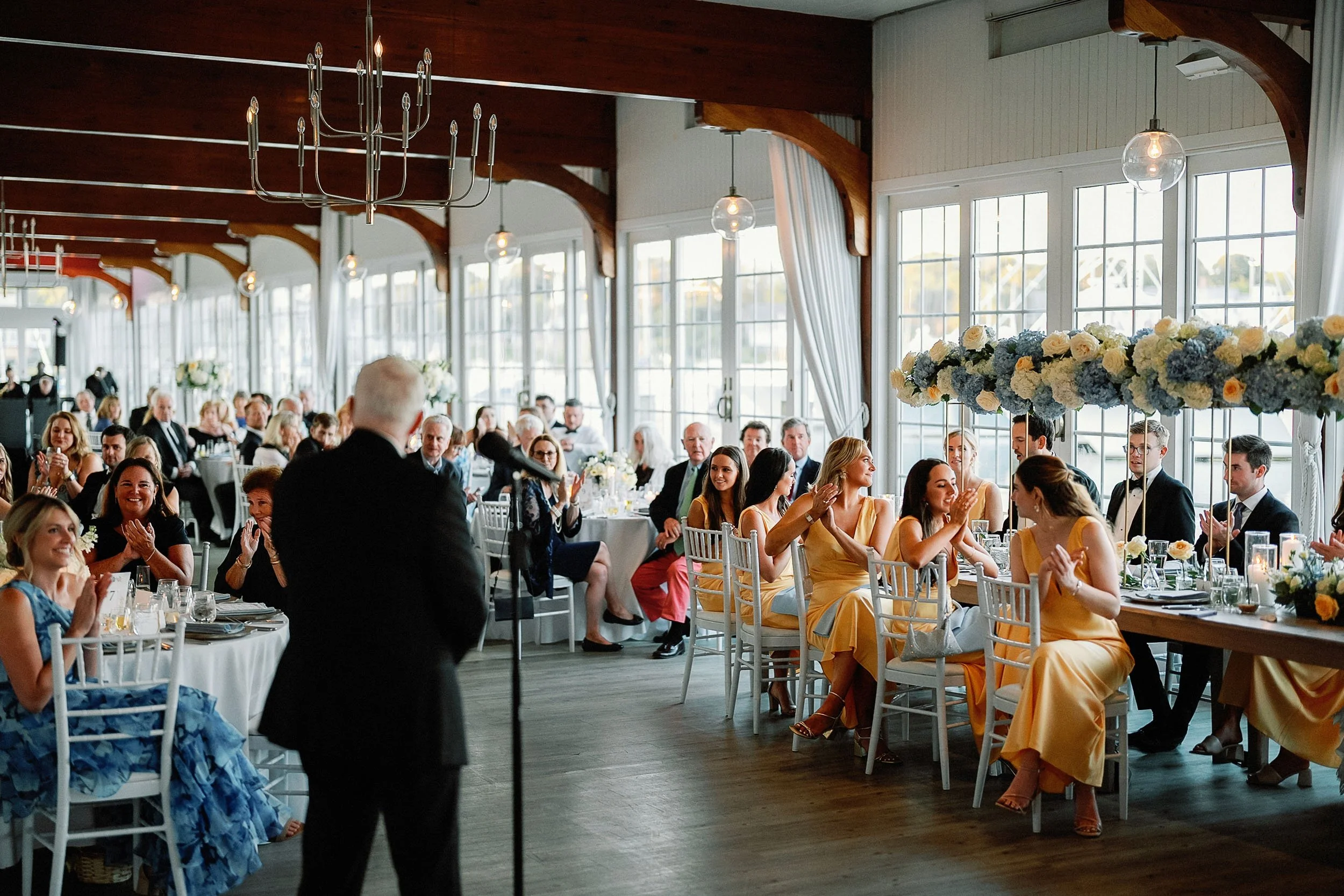 Guests seated at tables during a wedding reception, listening to a speaker, with floral centerpieces and large windows in a decorated event hall.