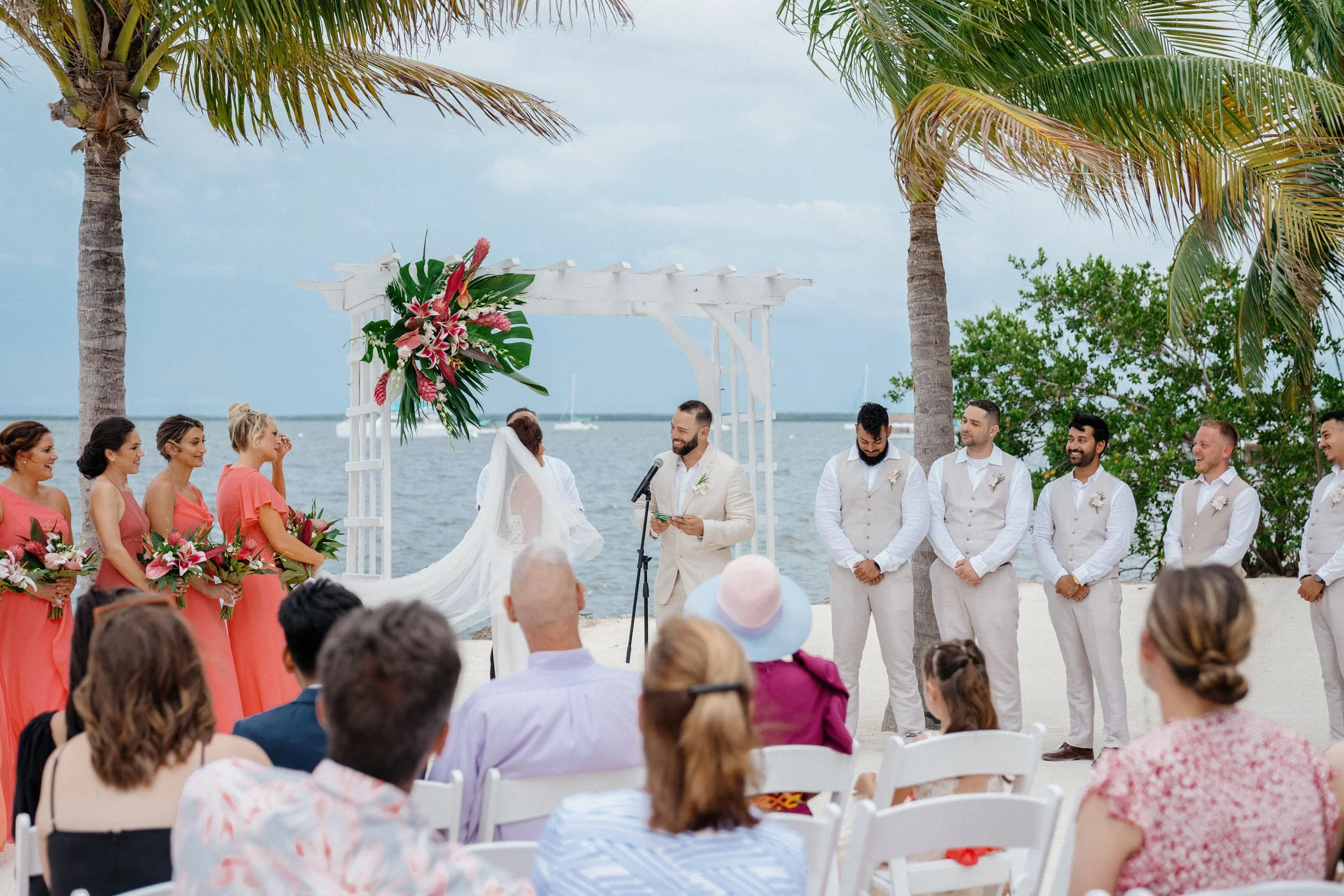 A wedding ceremony taking place on a beach with palm trees in the background, where a bride and groom are exchanging vows under a decorated arch with pink flowers, surrounded by bridesmaids in pink dresses and groomsmen in light suits, with guests se