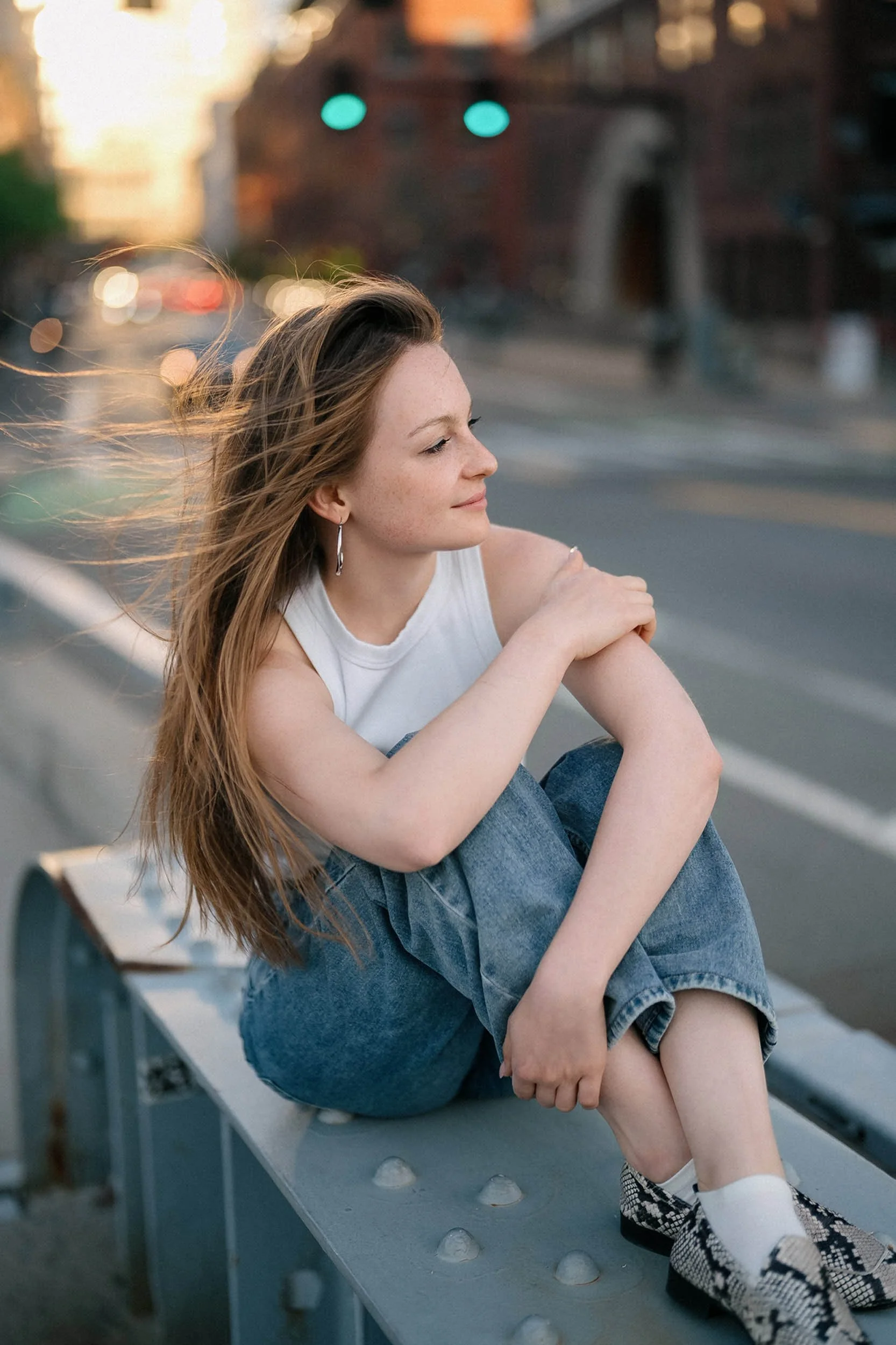 A young woman with long hair sitting on a metal barrier along a city street, with a sunset background and traffic lights in the distance.