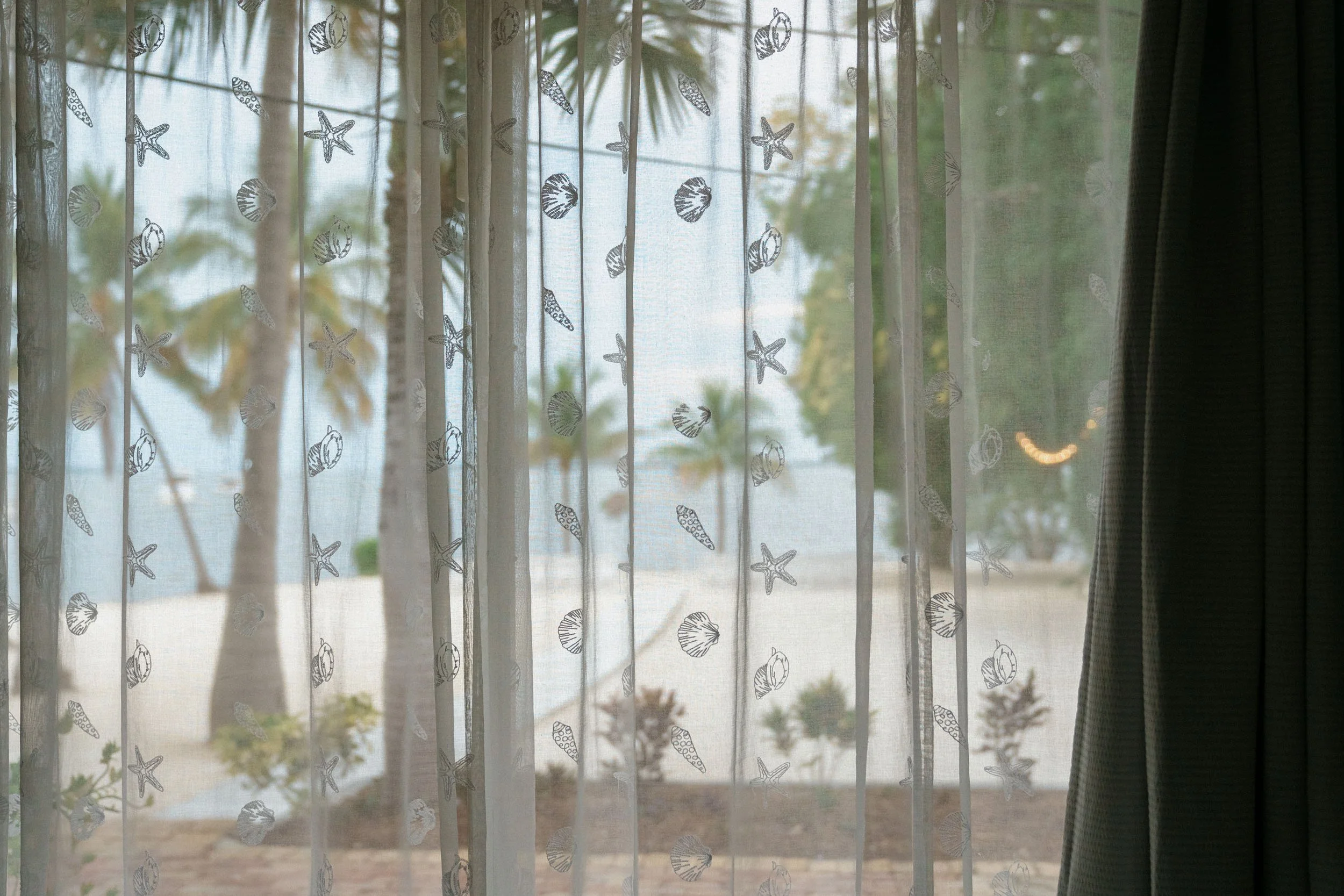 View through a sheer curtain with shell and starfish design, looking outside at palm trees, plants, and a sandy beach near the water.
