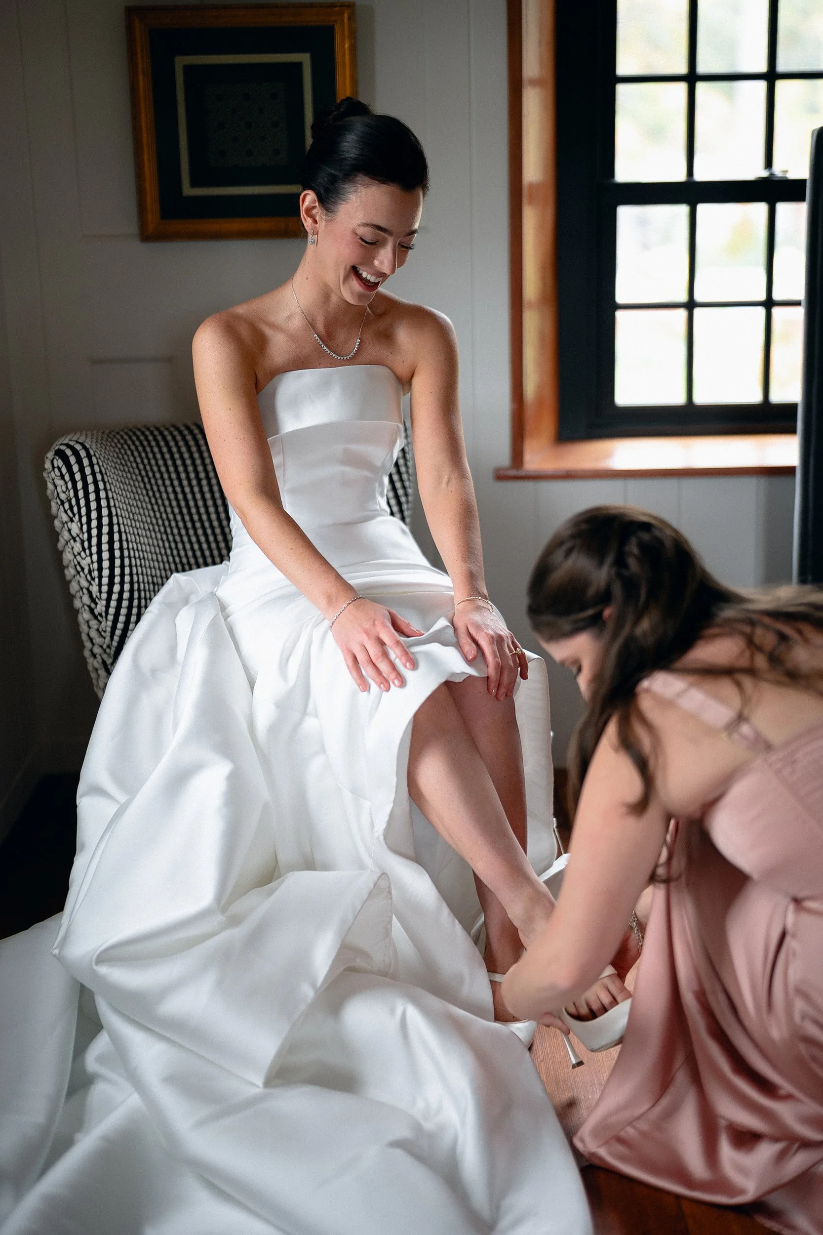 A bride in a strapless white wedding gown laughing as a woman helps her put on white shoes, in a room with a window and framed picture on the wall.