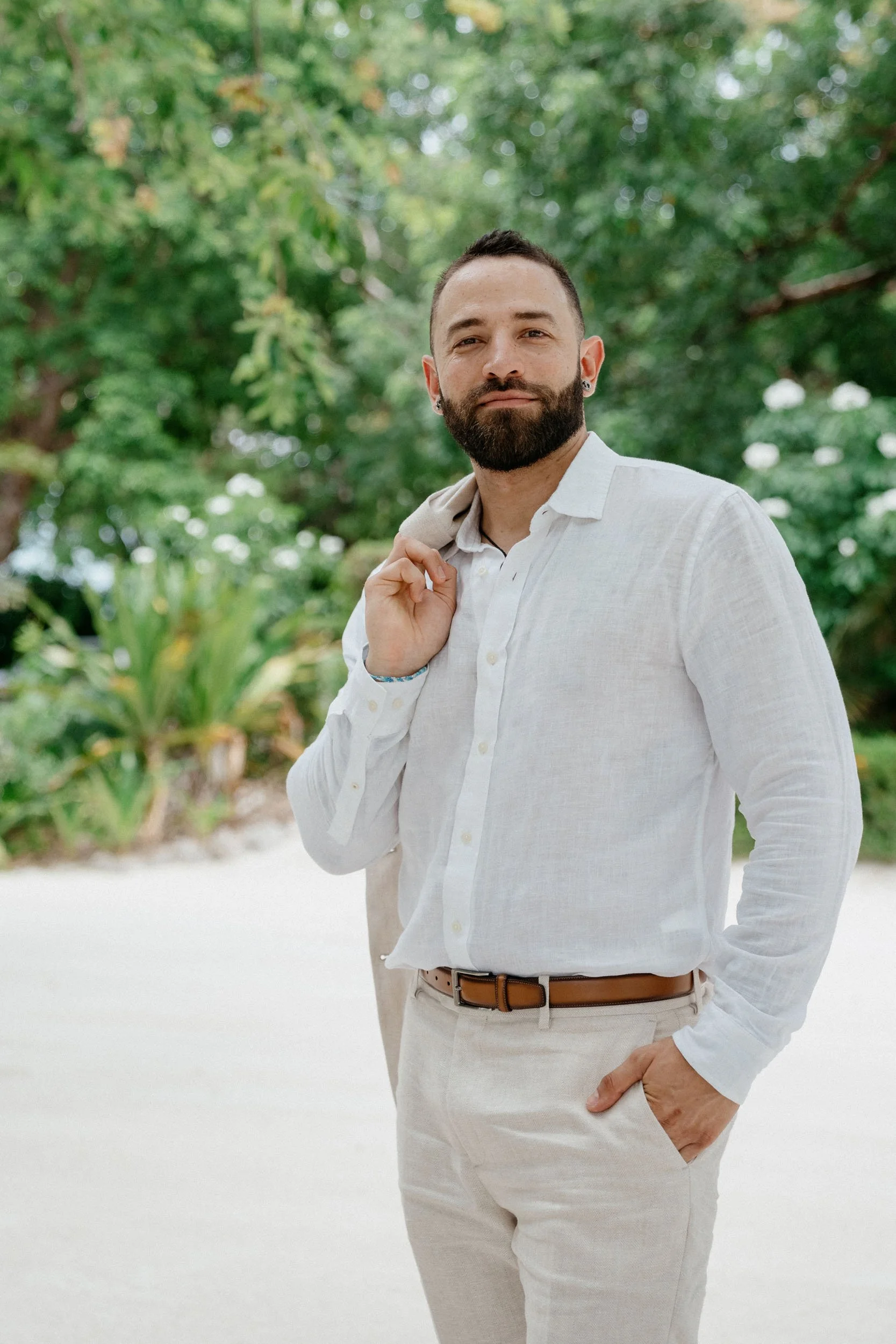A bearded man in a white linen shirt and beige pants standing outdoors with greenery in the background, holding a jacket over his shoulder.