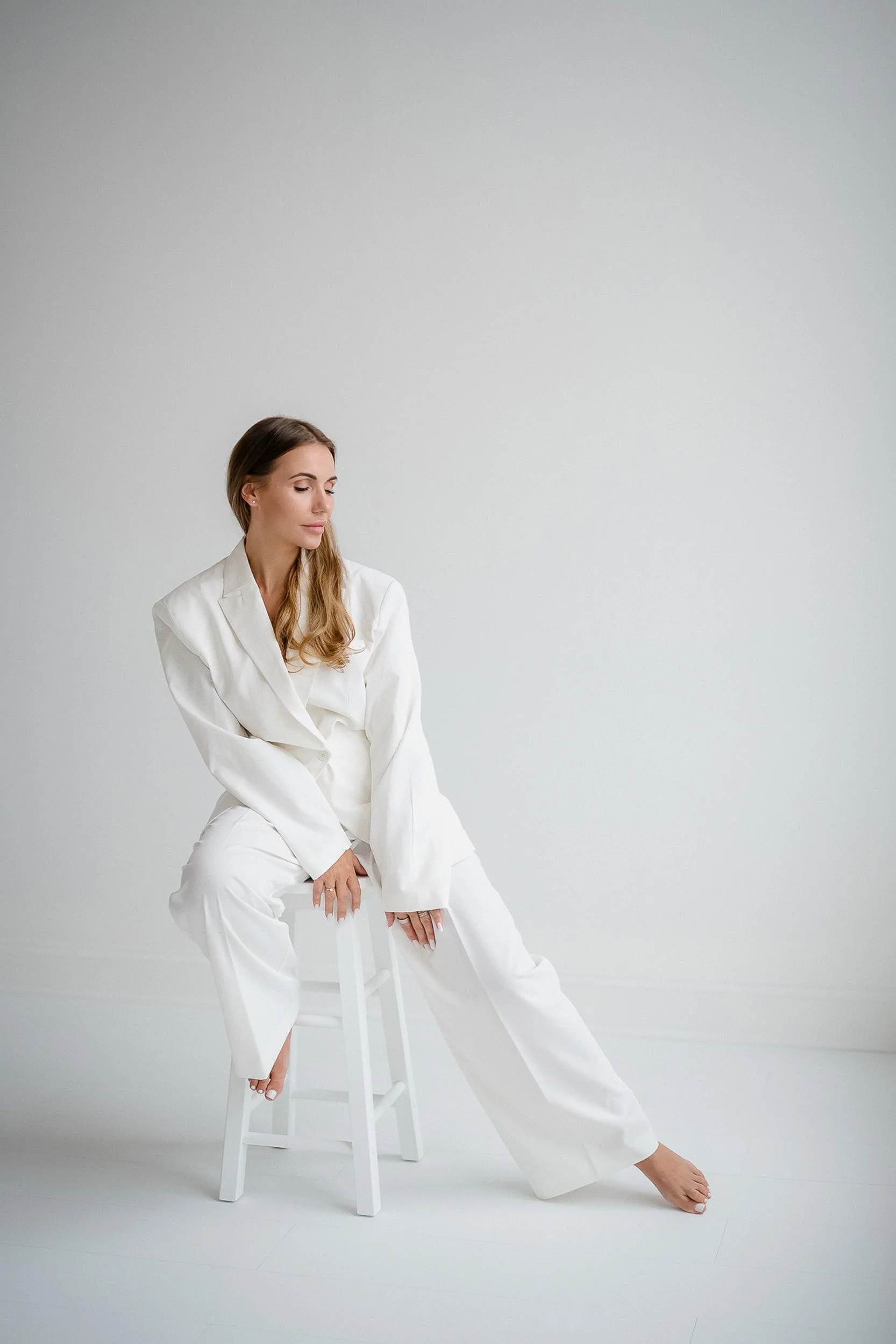A woman in a white suit sitting sideways on a white stool in a minimalist white room.