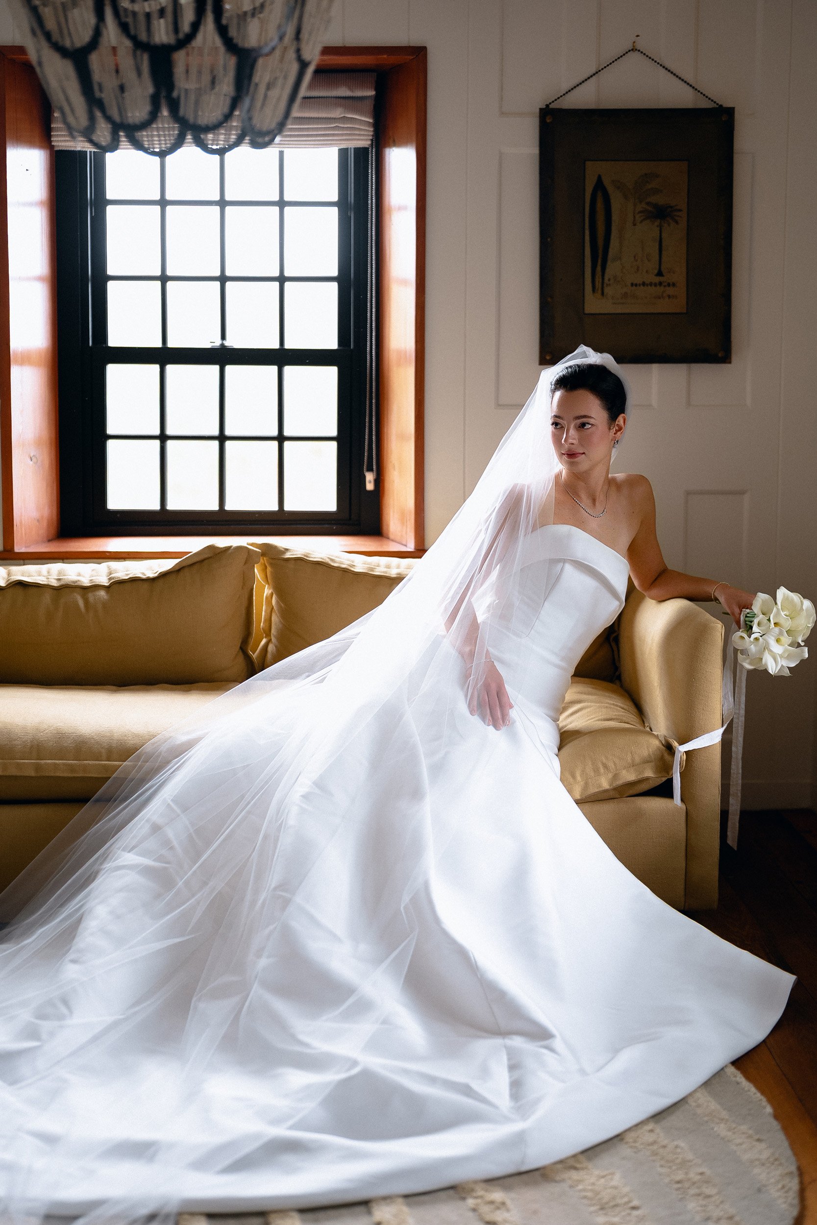 A bride in a white wedding dress with a veil, sitting on a yellow sofa in a room with a window and artwork on the wall.
