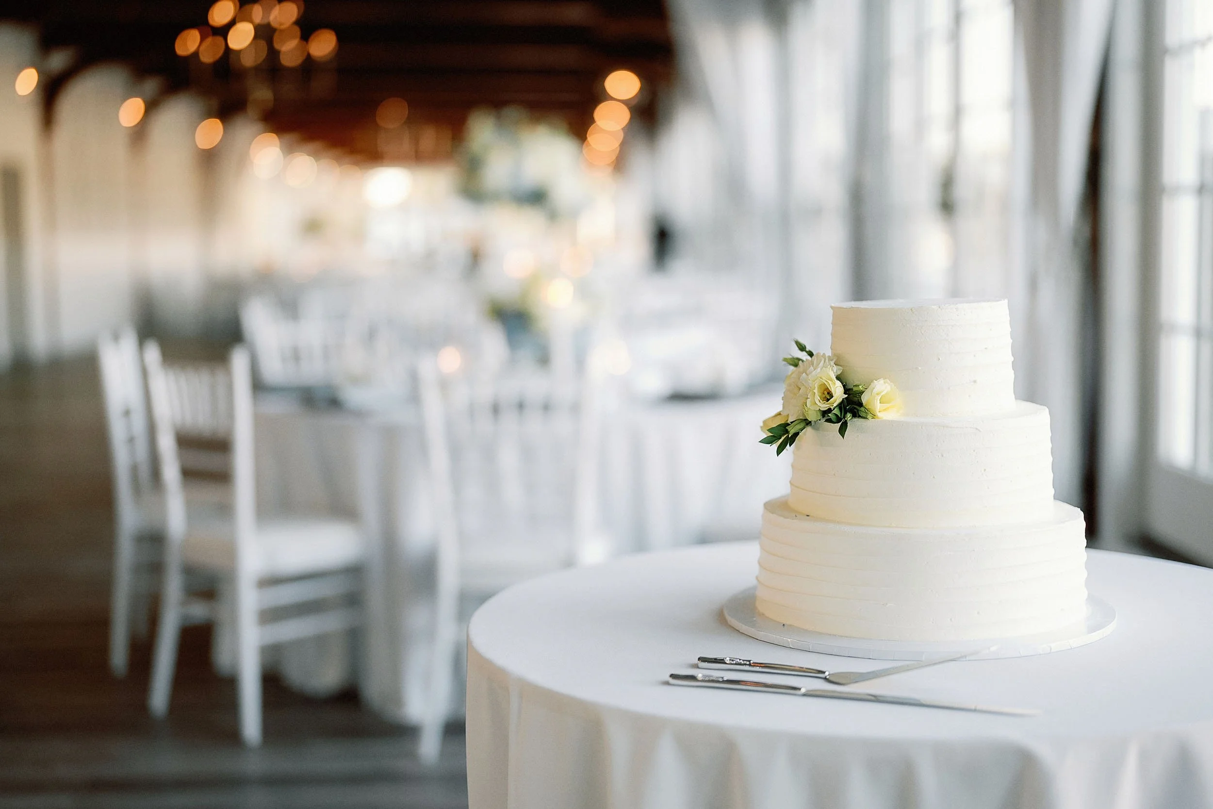 Three-tiered white wedding cake decorated with white roses and greenery, placed on a table with a white tablecloth in a softly lit reception hall.