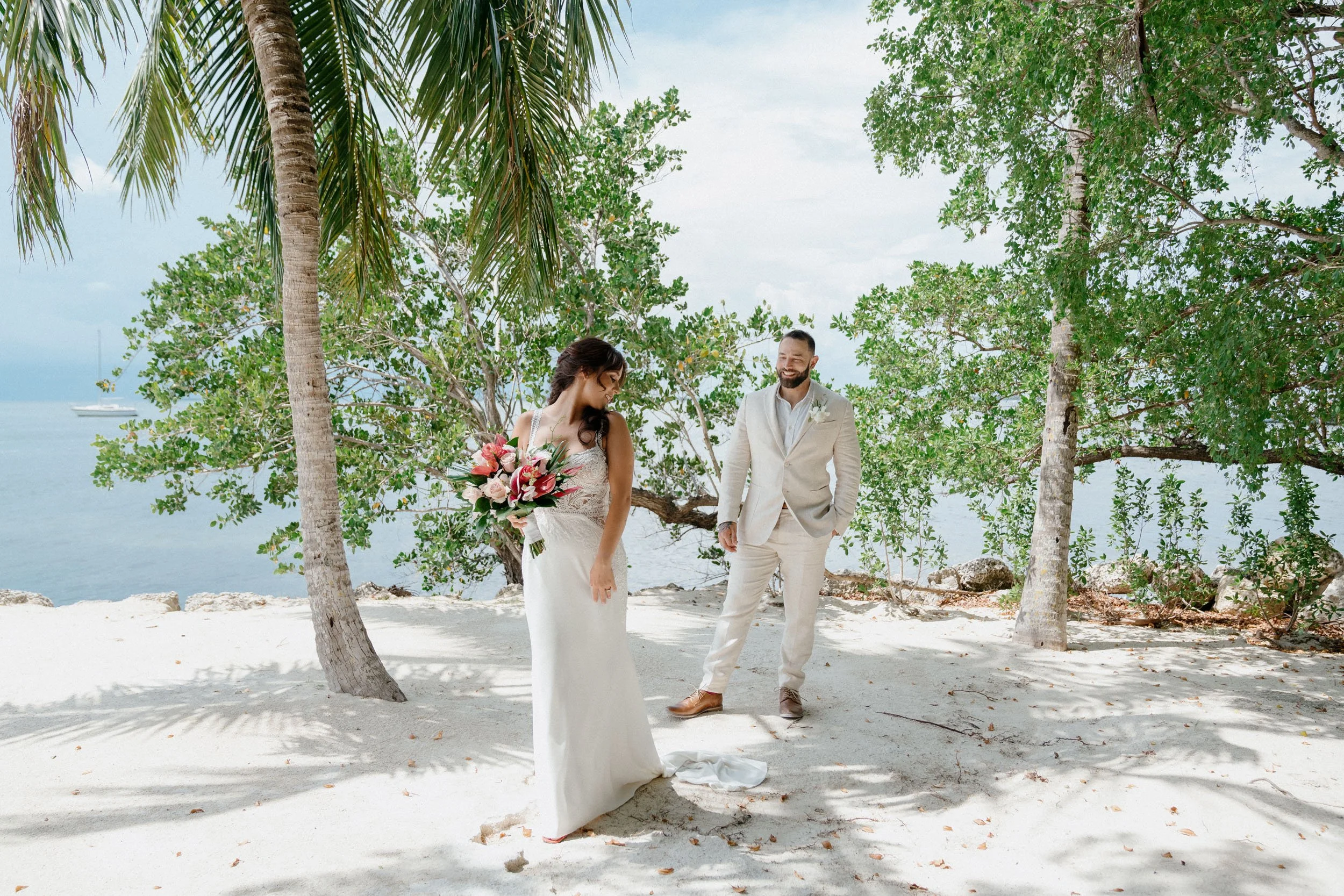 A bride and groom on a beach, with the bride holding a bouquet of flowers, surrounded by trees and overlooking the ocean.