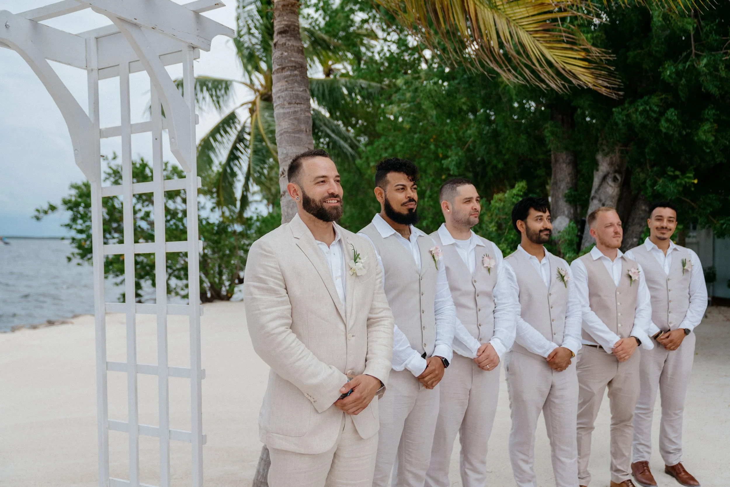Group of men dressed in light-colored suits standing on a beach, with trees and water in the background, during a wedding ceremony.