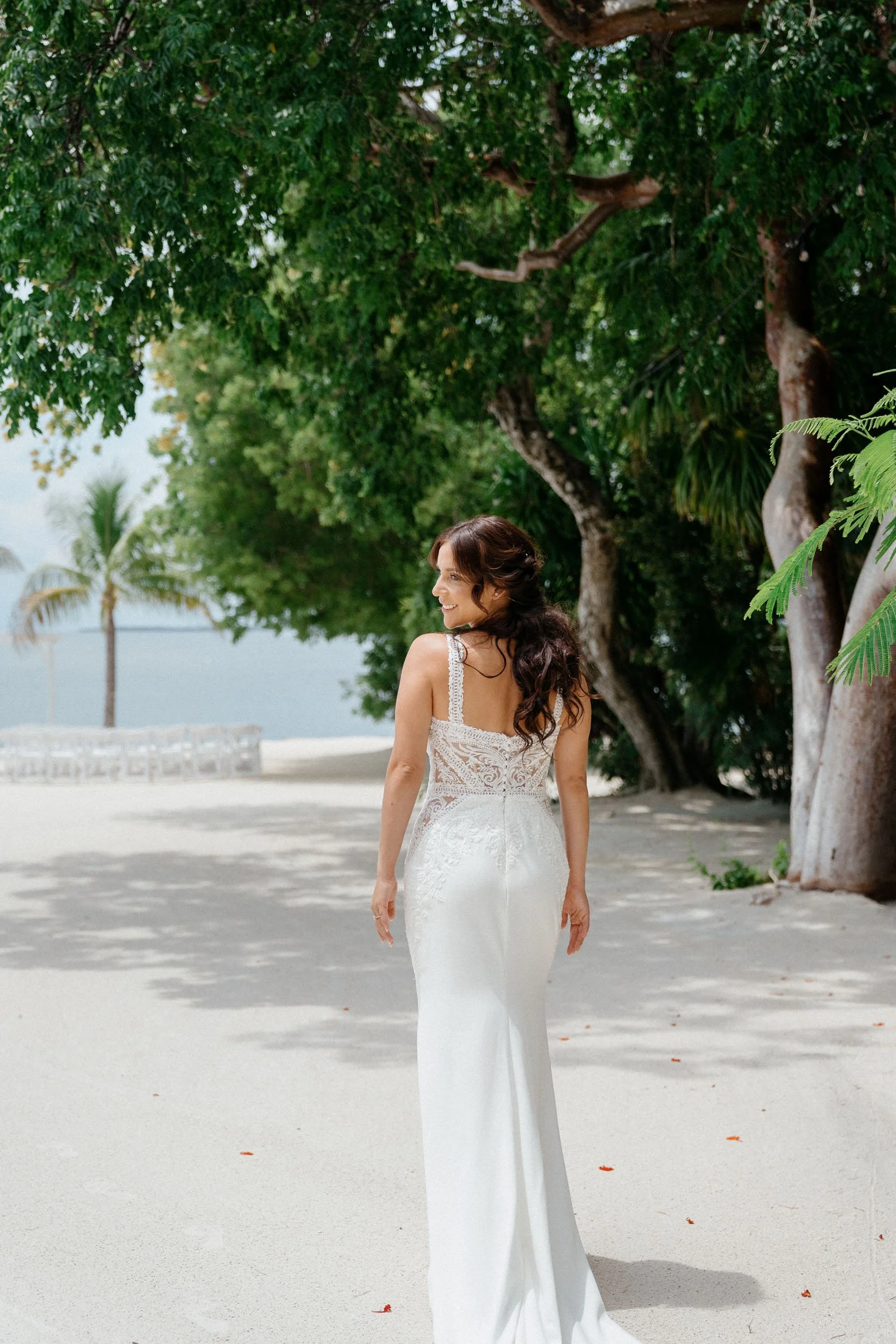 A woman in a white lace wedding dress walking on a sandy beach with trees and the ocean in the background, smiling and looking to her right.