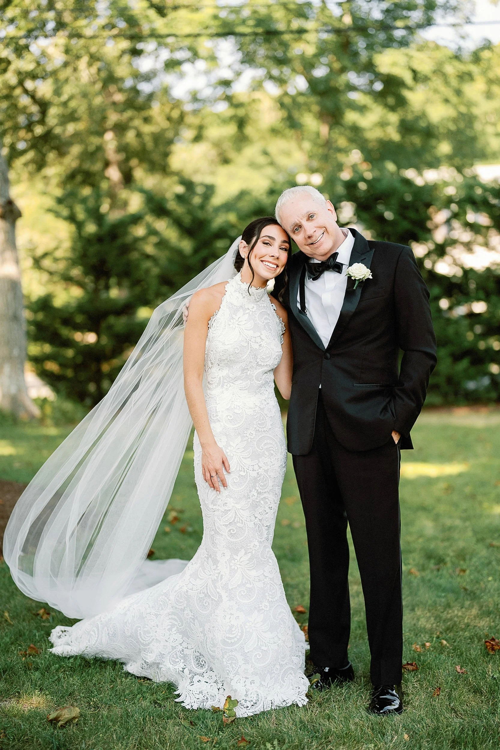 Bride in a white lace wedding gown and veil smiling with an older man in a black tuxedo outdoors surrounded by trees.