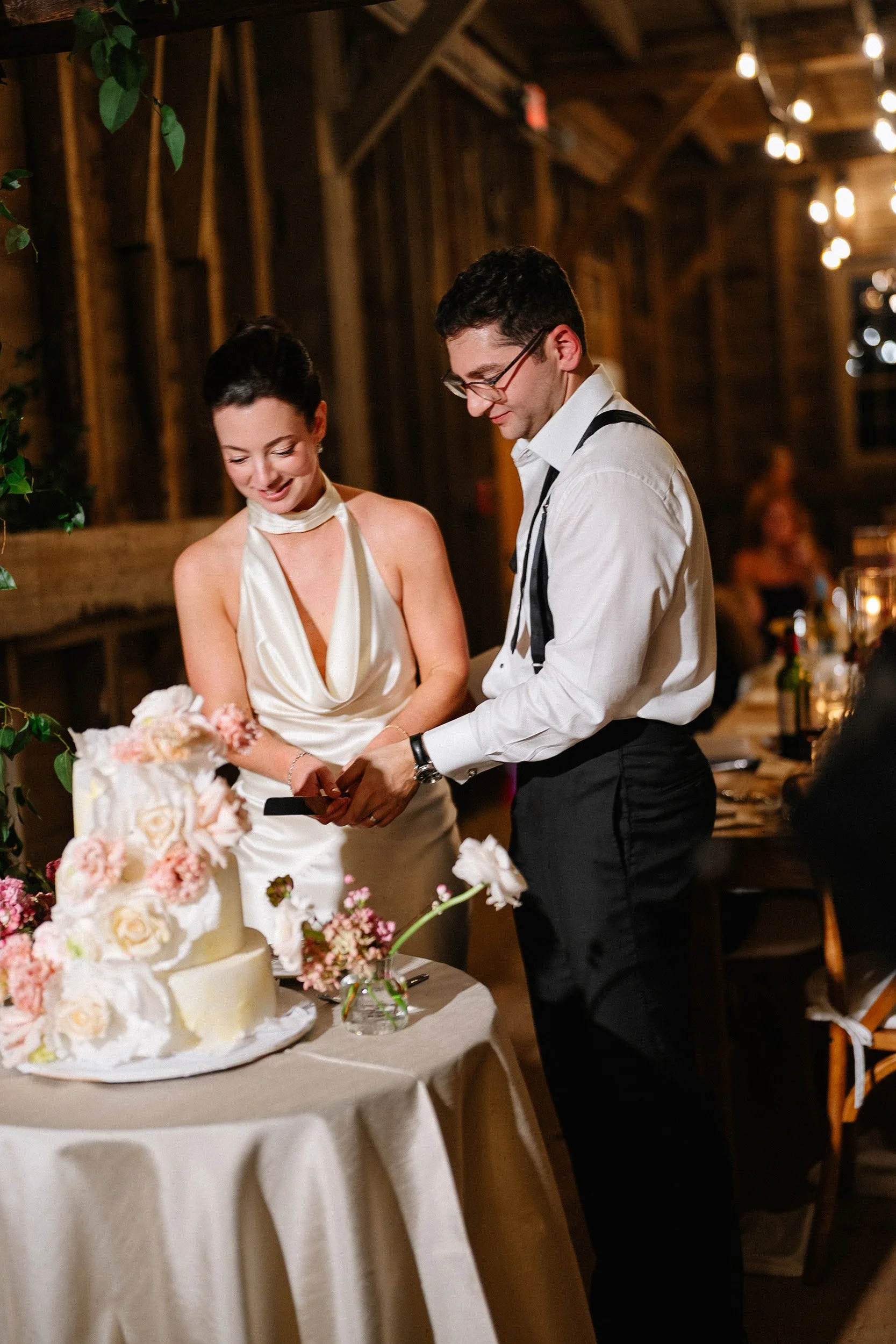 A bride and groom cutting a wedding cake together at the reception, both smiling, with the cake decorated with pink and white flowers.