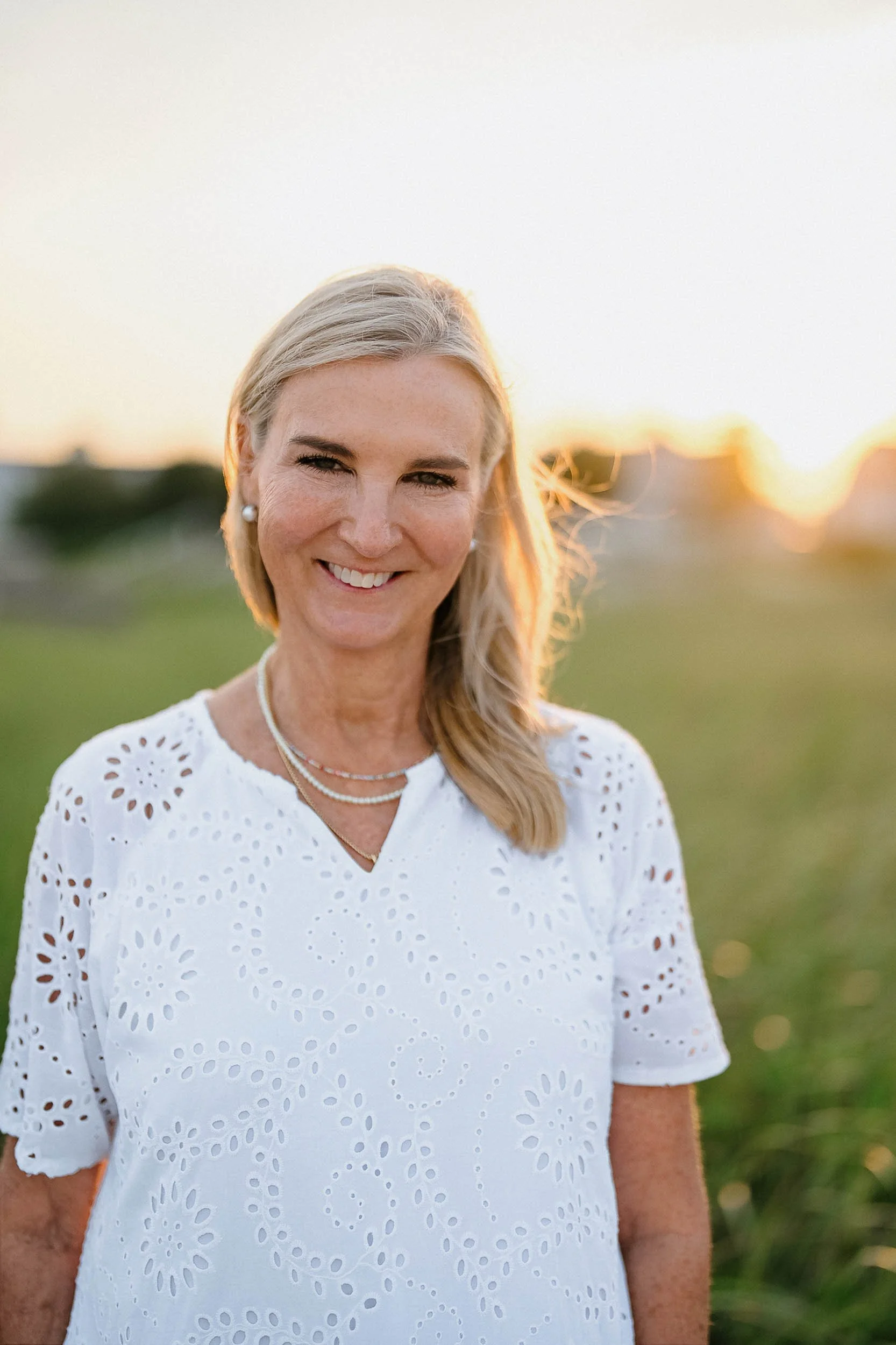 A woman with blonde hair smiling outdoors during sunset, wearing a white eyelet top and pearl jewelry.