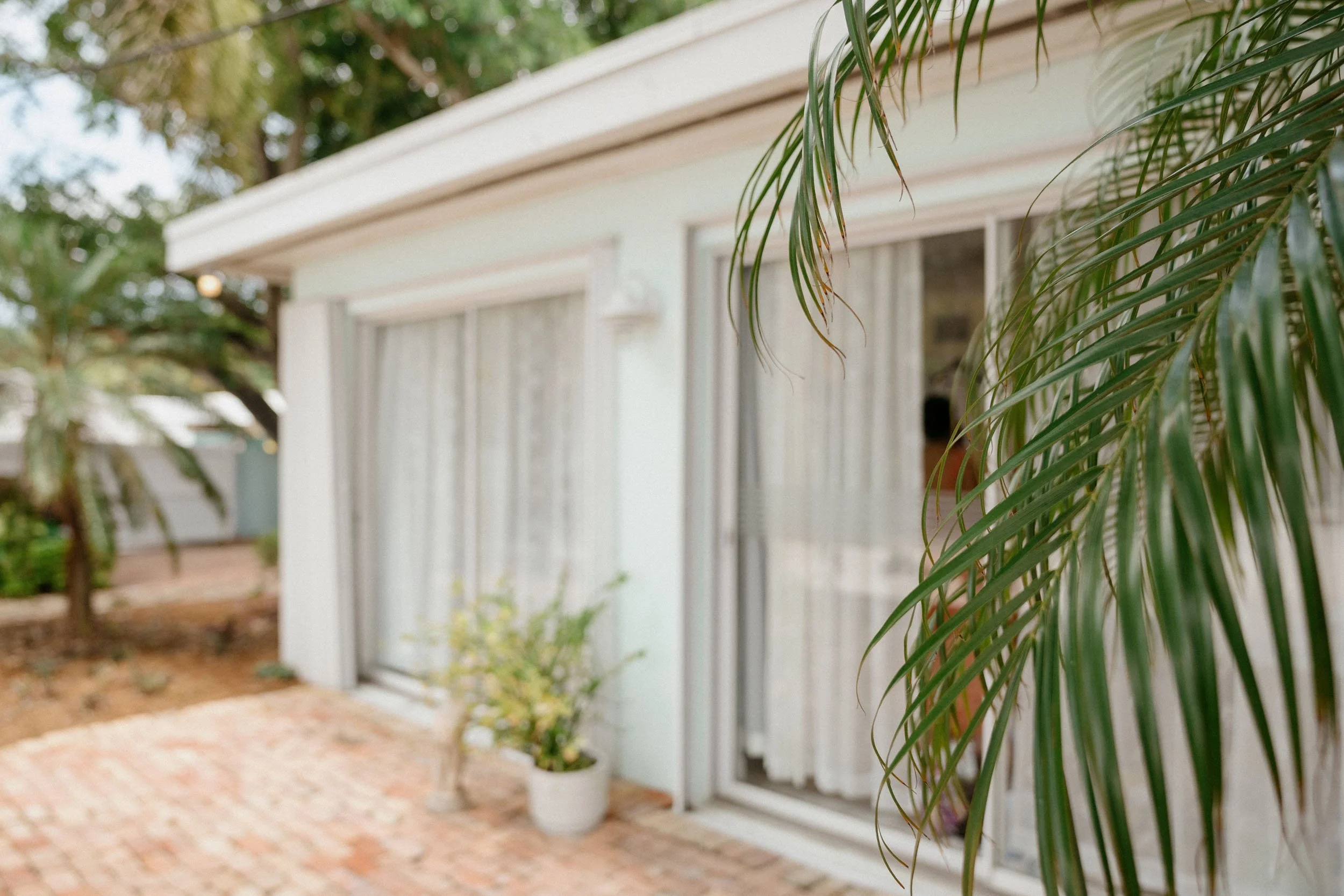 Exterior view of a house with sliding glass doors, white curtains, and potted plants, surrounded by greenery and brick flooring.