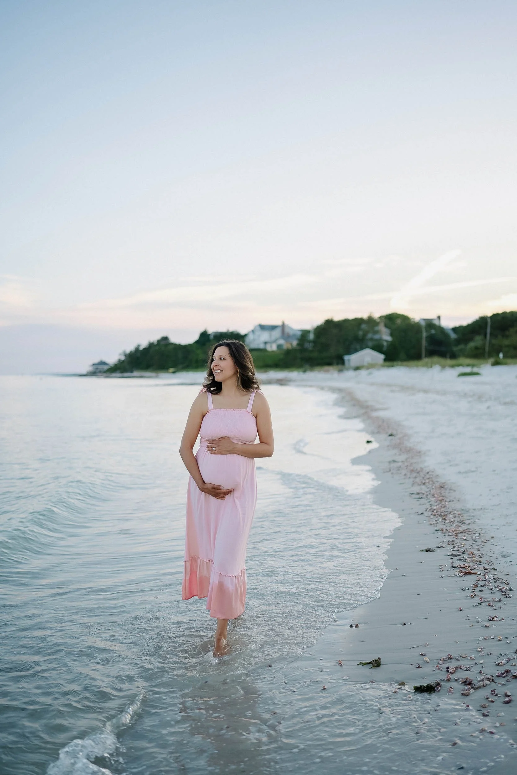 Pregnant woman in pink dress walking in shallow water on beach, smiling, with houses on a green hillside in the background during sunset.
