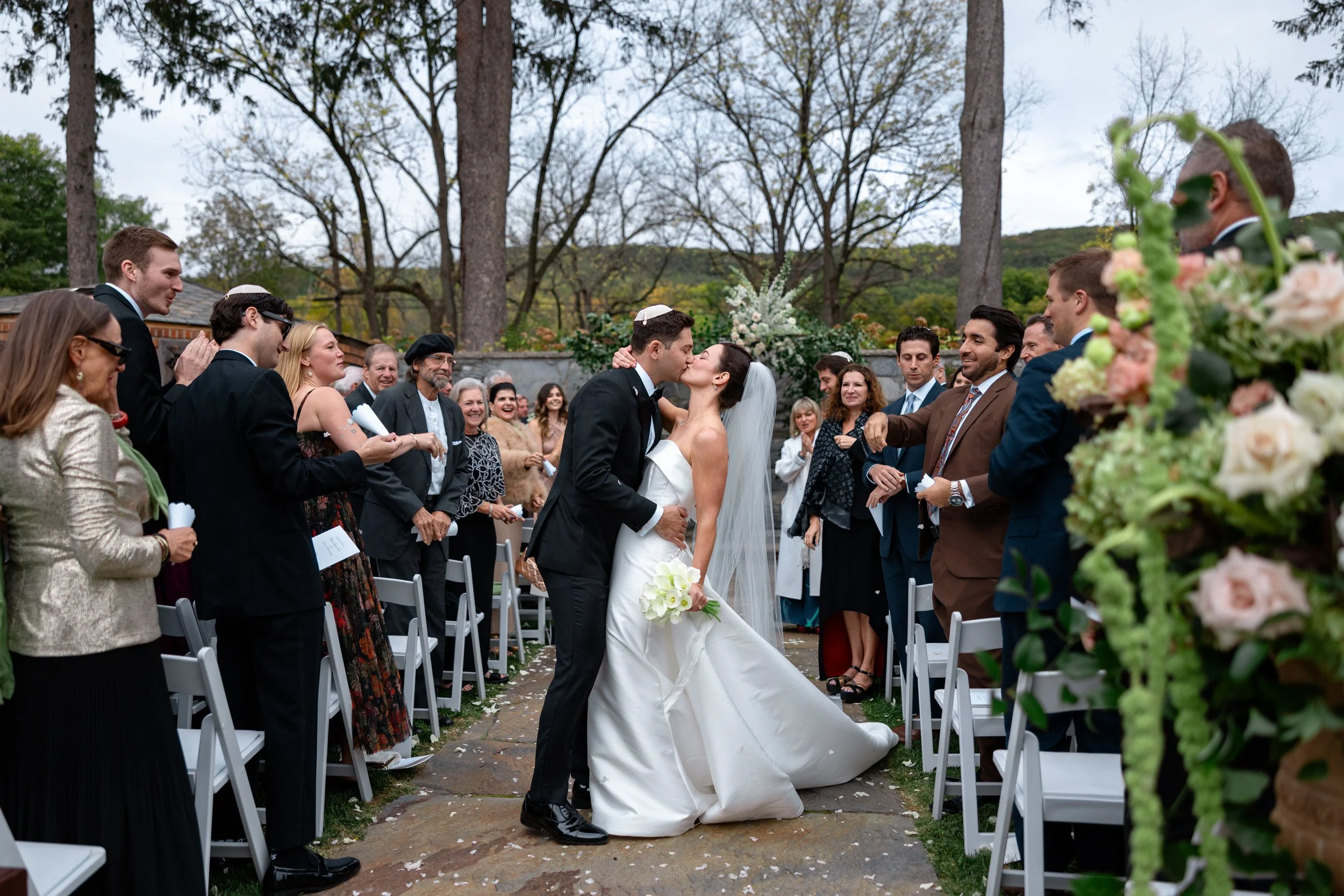 Bride and groom kissing during outdoor wedding ceremony with guests standing and sitting around them, trees in the background, and floral decorations.