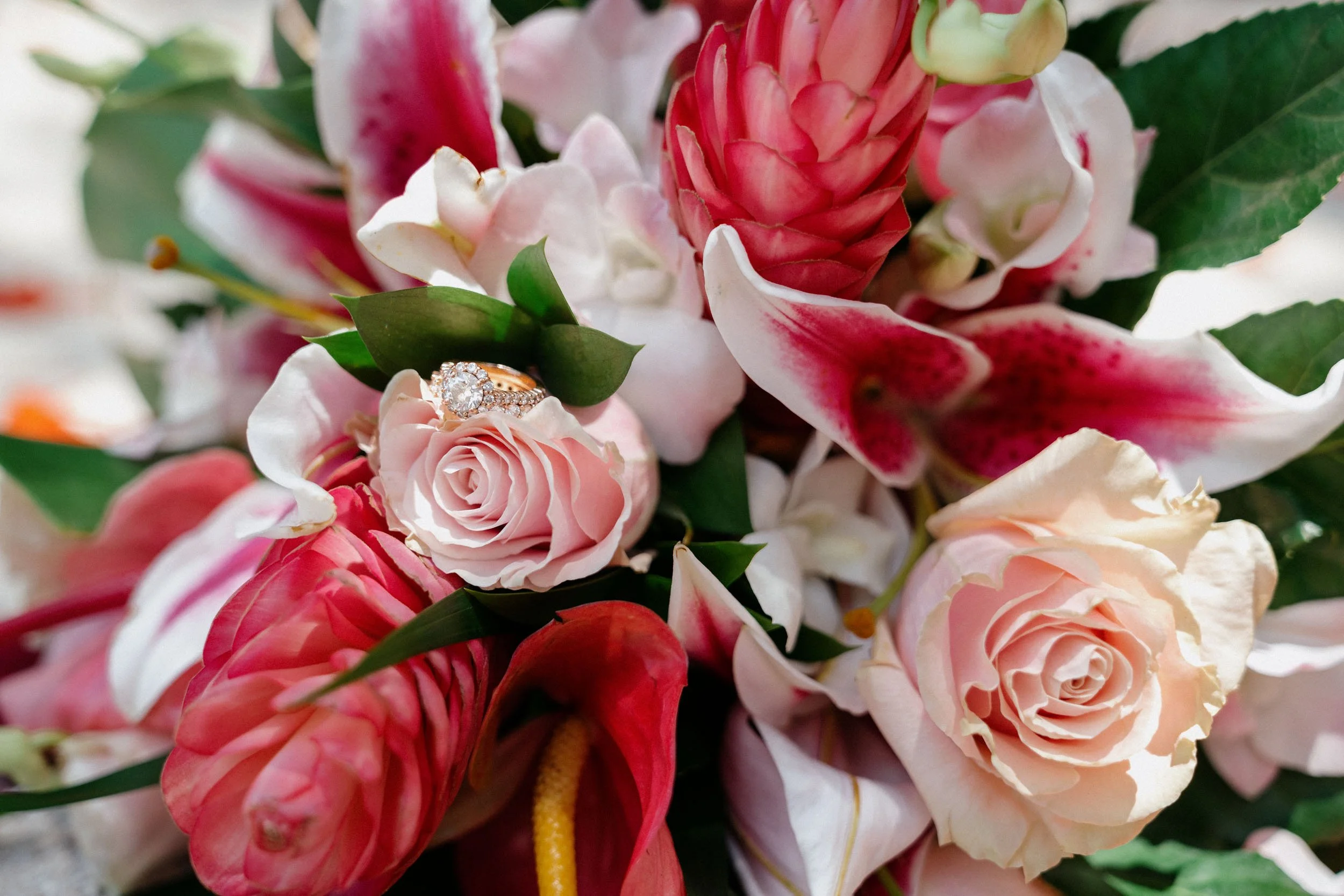 A close-up of a floral arrangement with pink roses, white and pink lilies, and green leaves. There is a wedding ring with diamonds resting inside a pink flower.