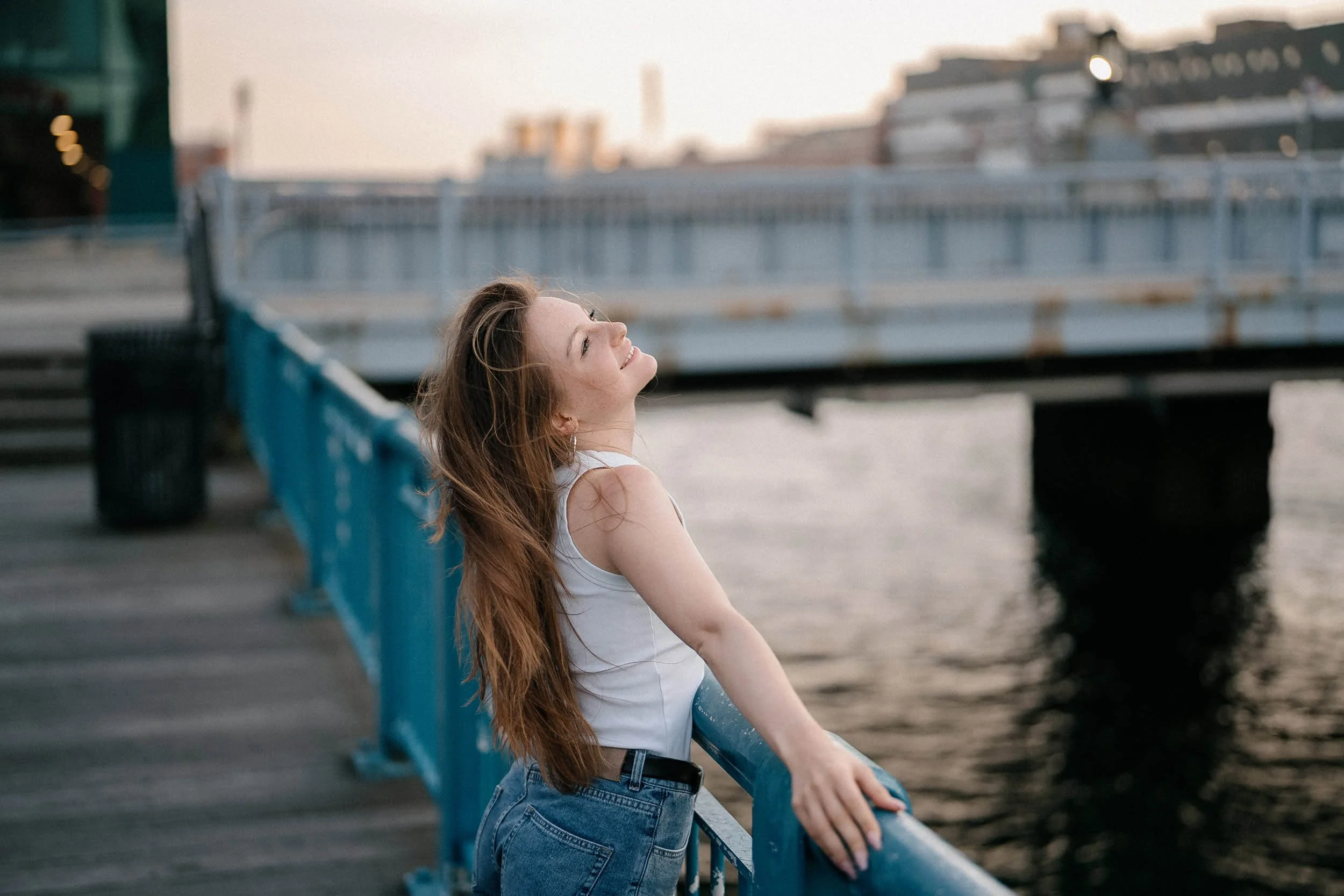 A young woman with long red hair smiling and leaning on a blue railing near a body of water, with a bridge and cityscape in the background during sunset.