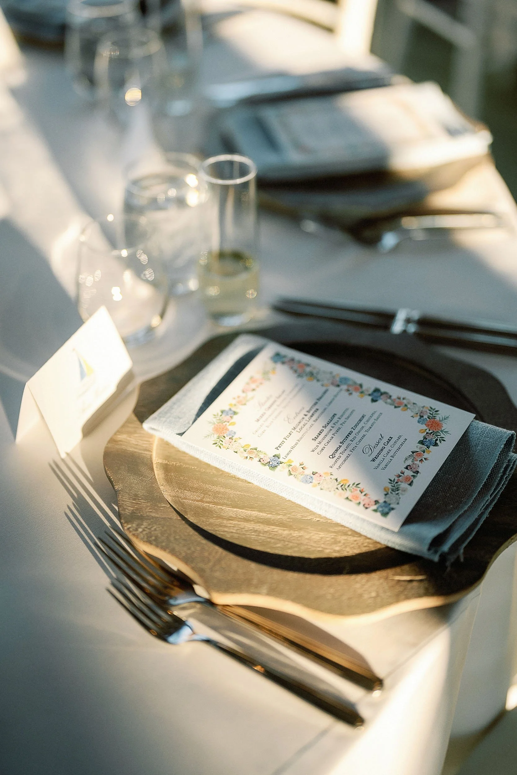 Elegant outdoor dining table setup with a white tablecloth, a wooden charger plate, a folded grey napkin, a printed menu with floral border, and glassware including a water glass and champagne flute.