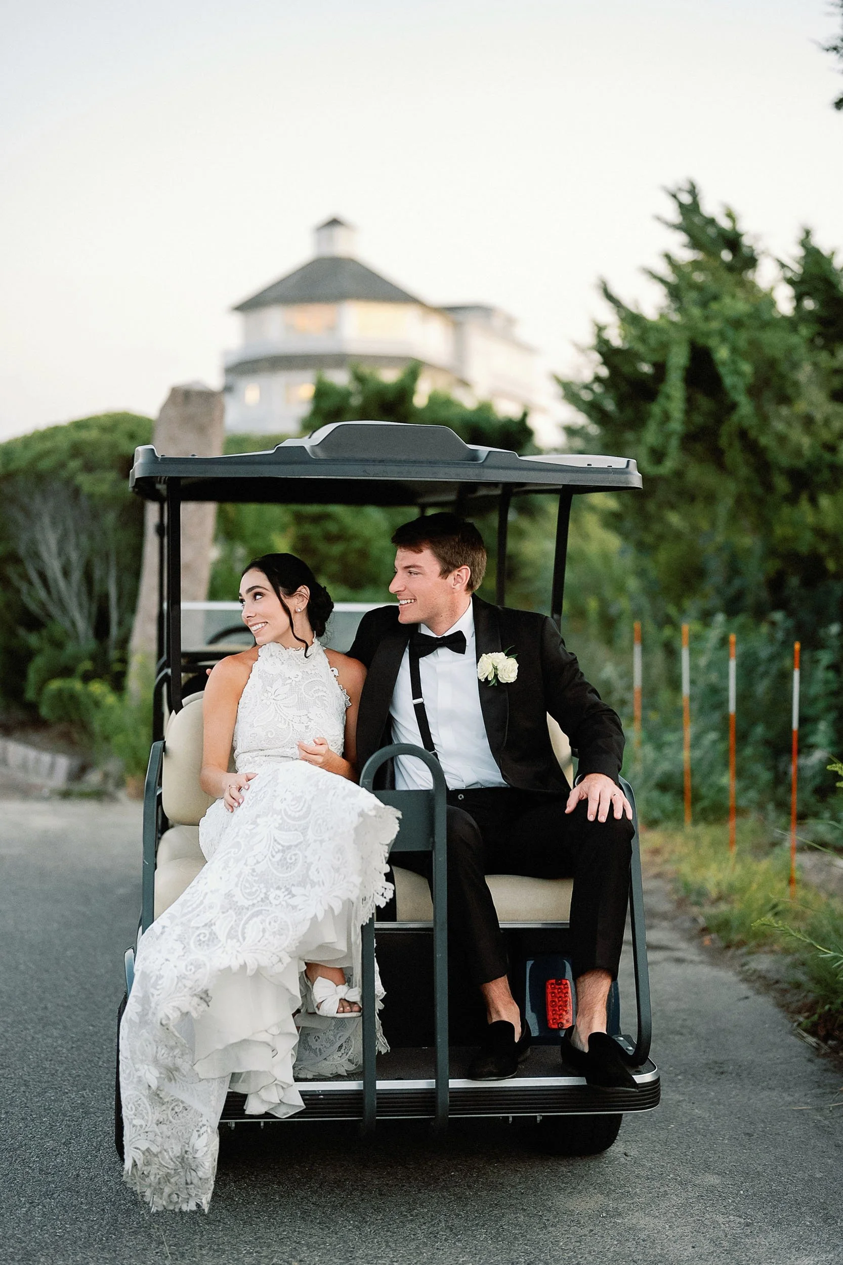 A newlywed couple in wedding attire sitting on a golf cart, smiling at each other, with a house and trees in the background during daytime.