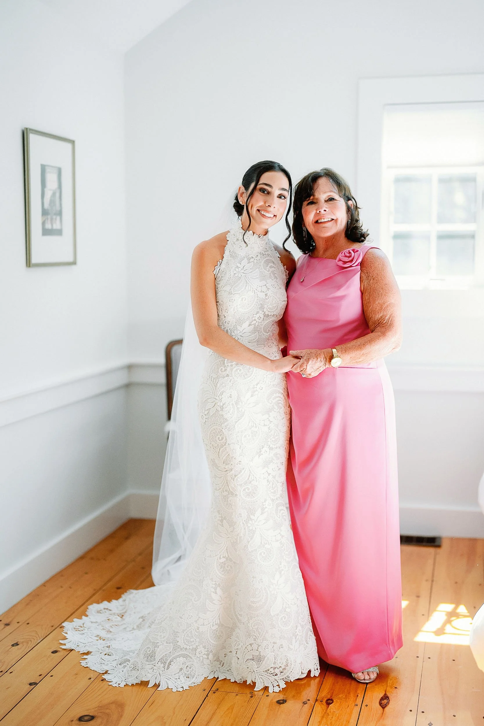 A bride in a lace wedding gown and a woman in a pink dress holding hands and smiling in a room with wooden floor and white walls.