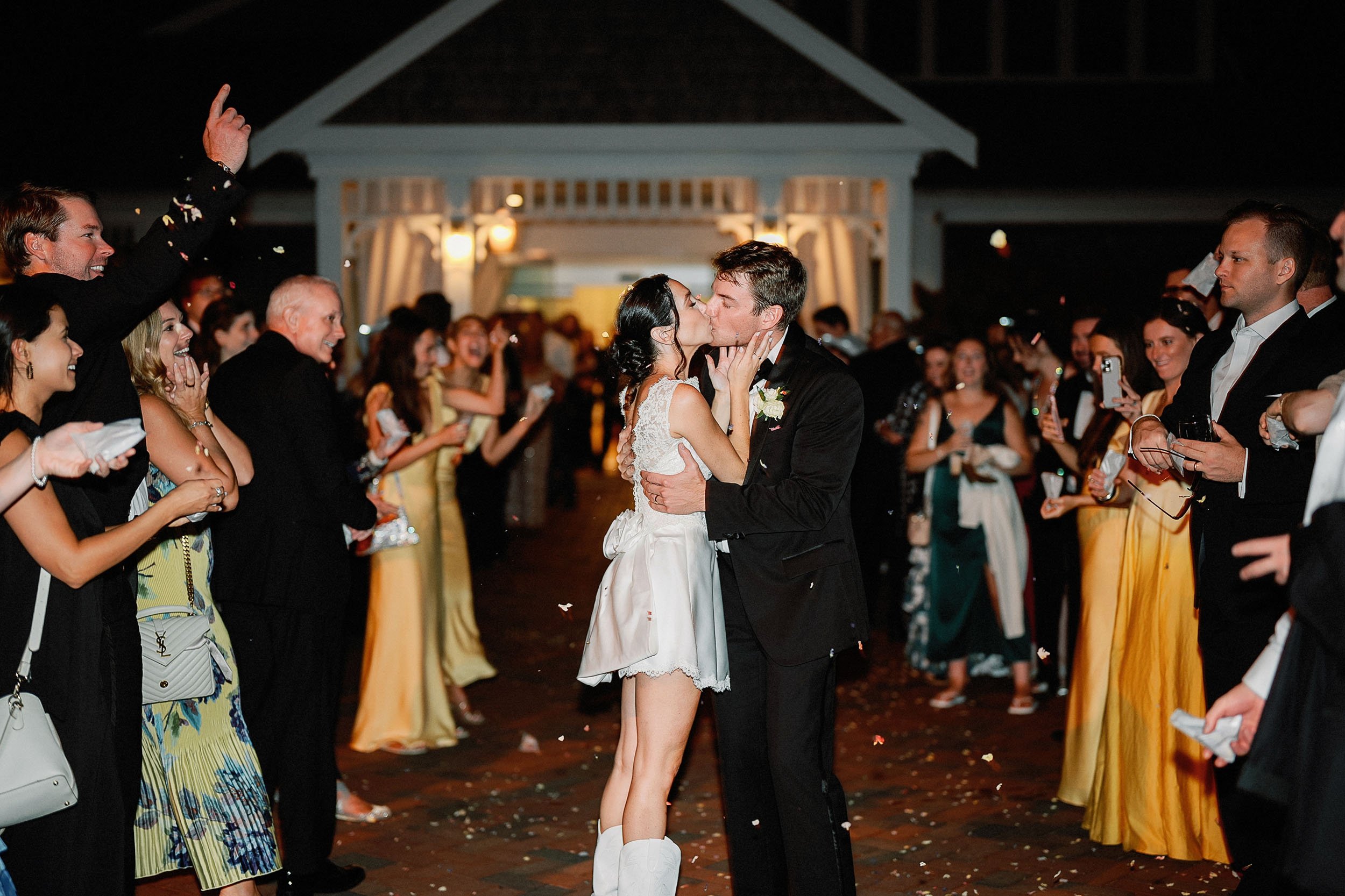 Couple kissing at their wedding reception, surrounded by friends and family, with some guests taking photos and celebratory confetti on the ground.